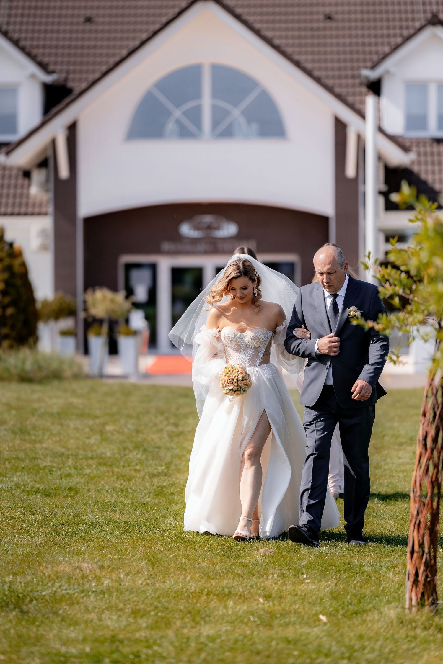 Sunny outdoor wedding scene with elegant bridal attire on landscaped lawn in front of white house.