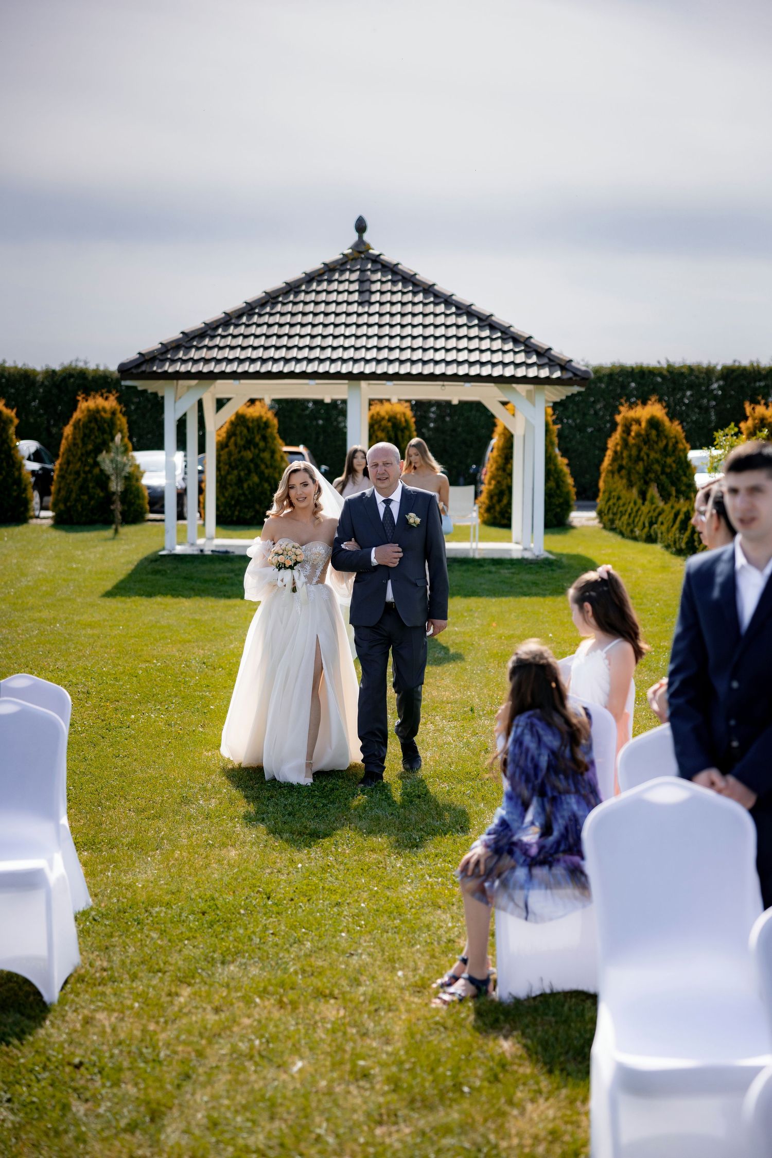 Outdoor wedding ceremony setup with white gazebo and chairs on manicured lawn surrounded by evergreen trees.