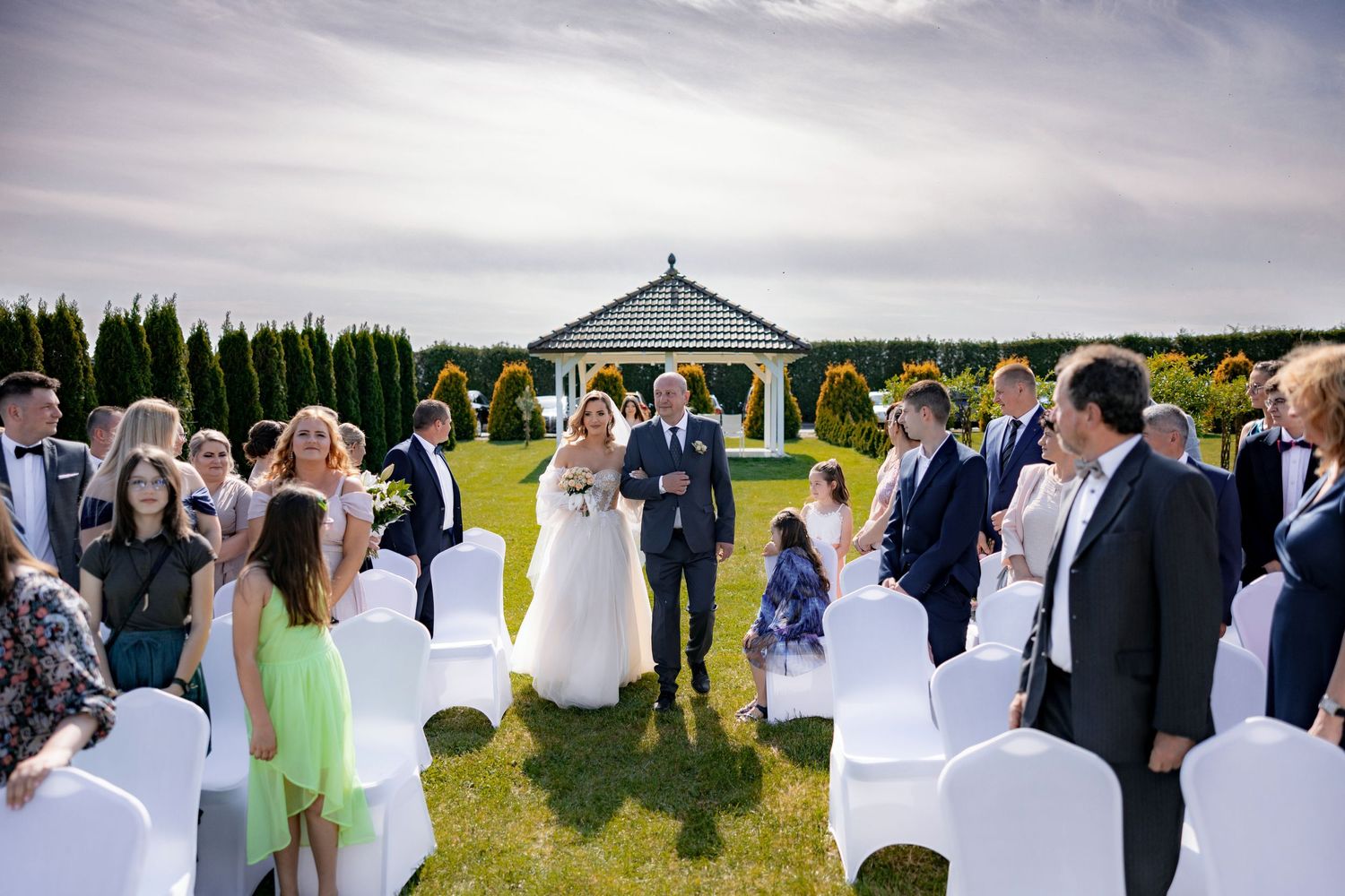 Outdoor garden wedding ceremony with white chairs and gazebo surrounded by guests.