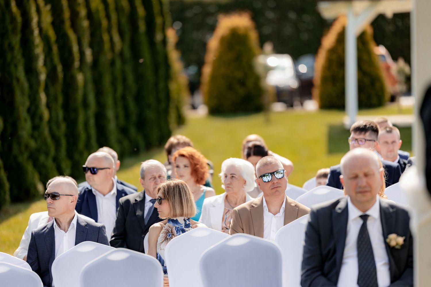 Wedding guests seated in white chairs during outdoor ceremony with manicured cypress trees.