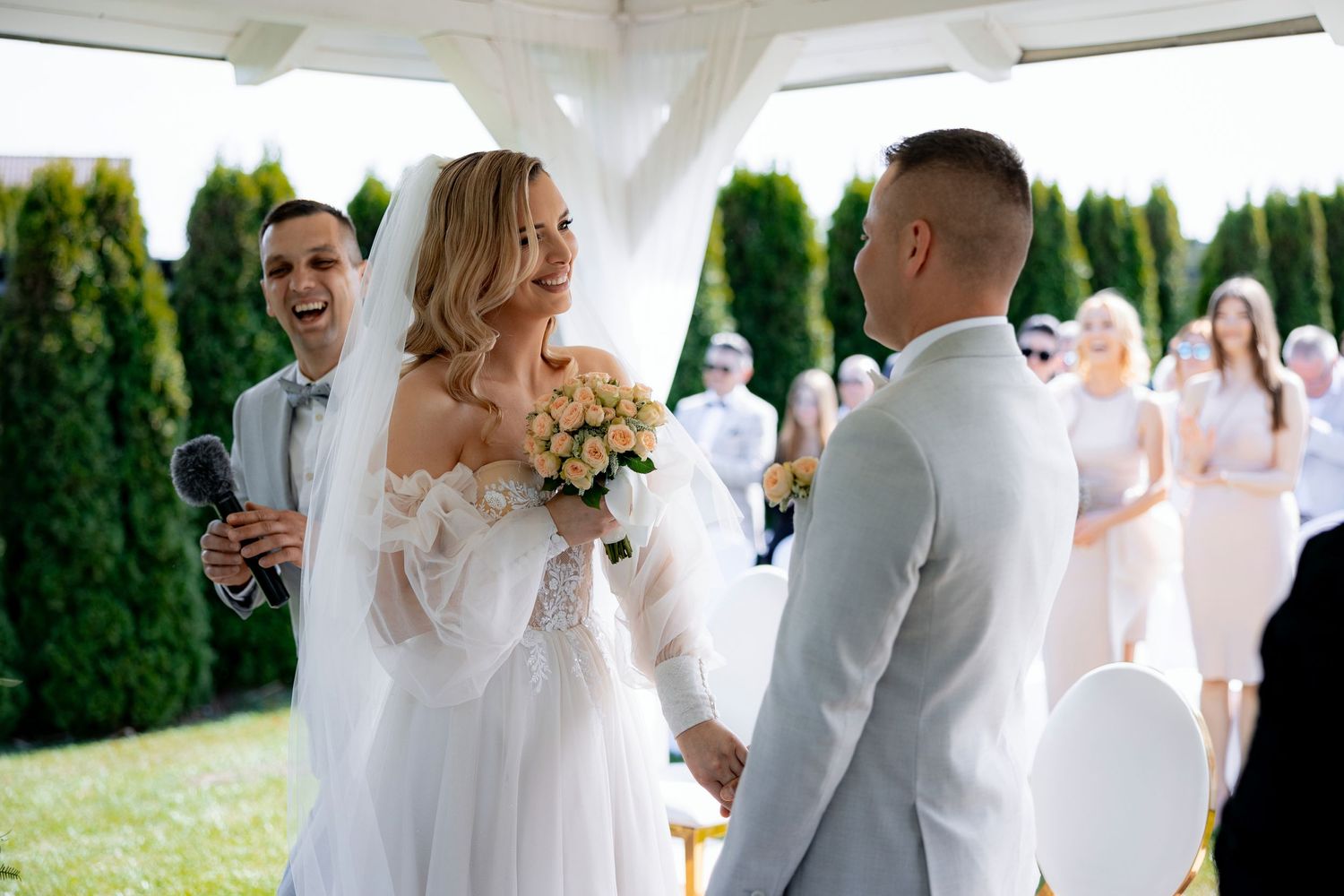 A joyful moment captured during an outdoor wedding ceremony under a white canopy with evergreen trees in the background.