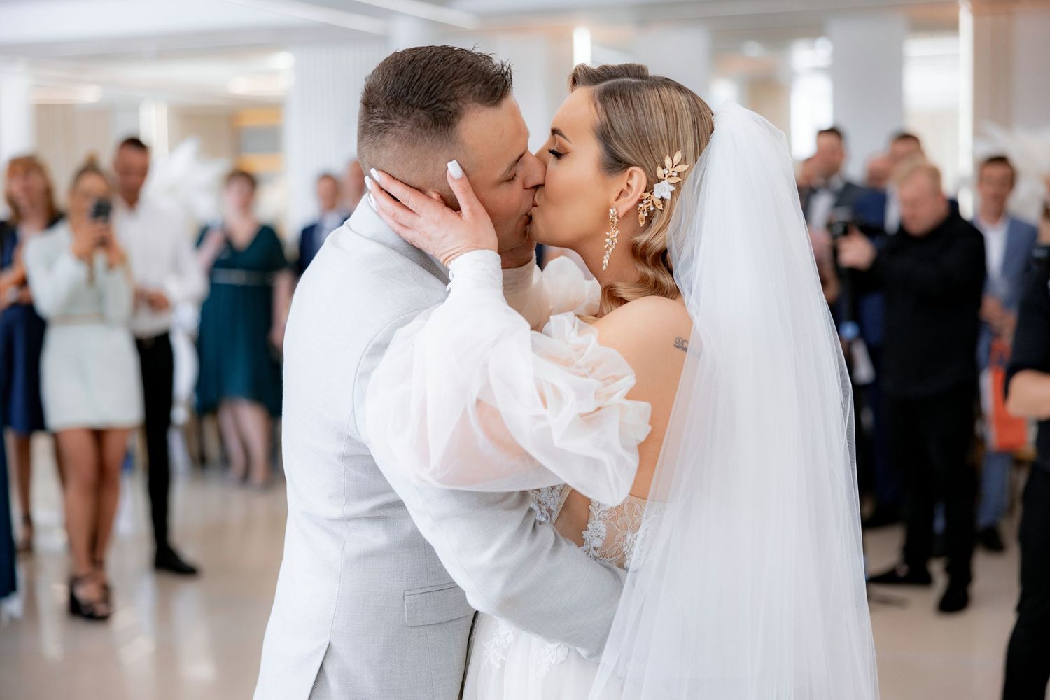 A tender kiss shared between newlyweds in white and gray attire as wedding guests look on in the background.