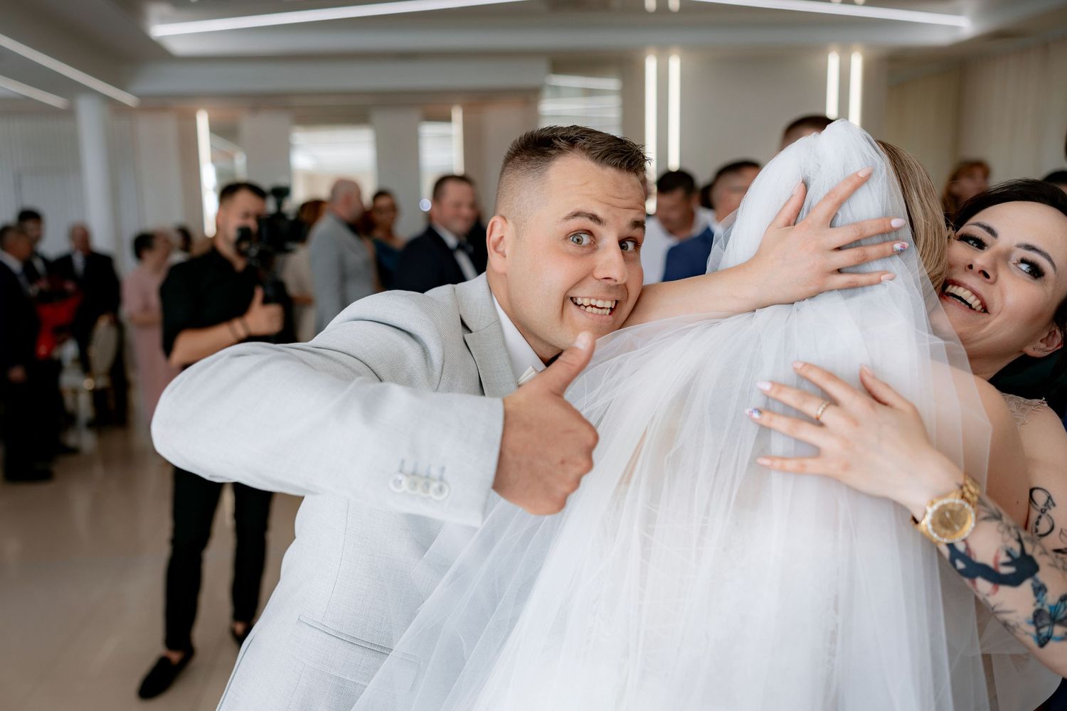 Playful moment captured as a groom in light grey suit lifts a flowing white veil.