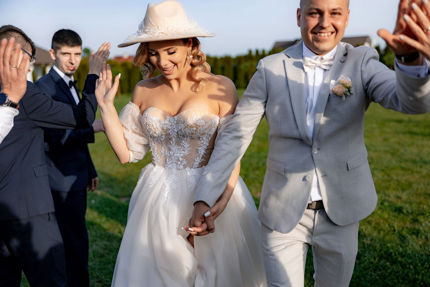 Newlyweds celebrate joyfully outdoors with guests as bride wears flowing white gown and straw hat.
