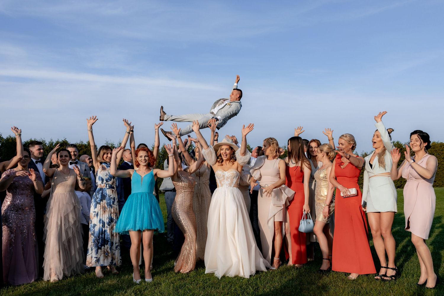 Wedding guests toss groom in air during outdoor celebration.