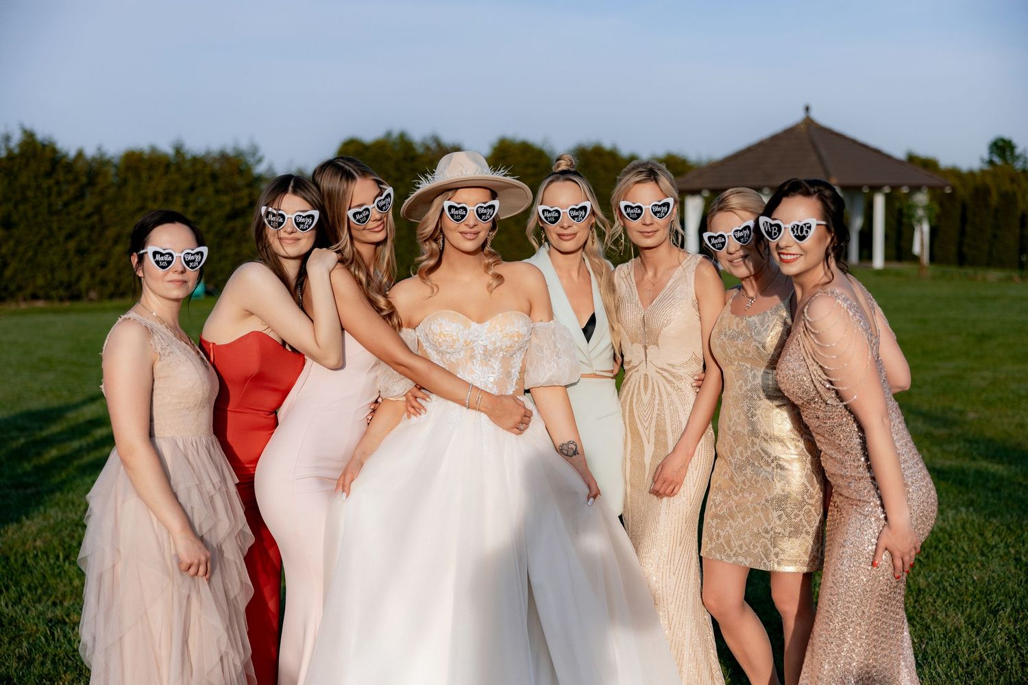 Bridesmaids in pastel dresses and sunglasses pose with the bride on a sunny lawn.
