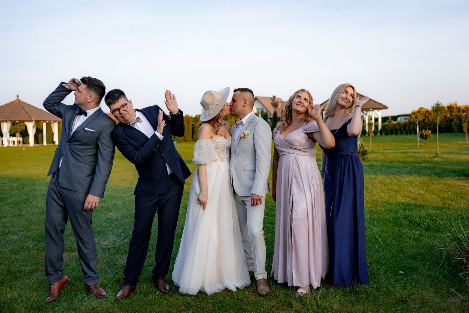 Wedding party poses playfully on a lawn at sunset with a gazebo in background.