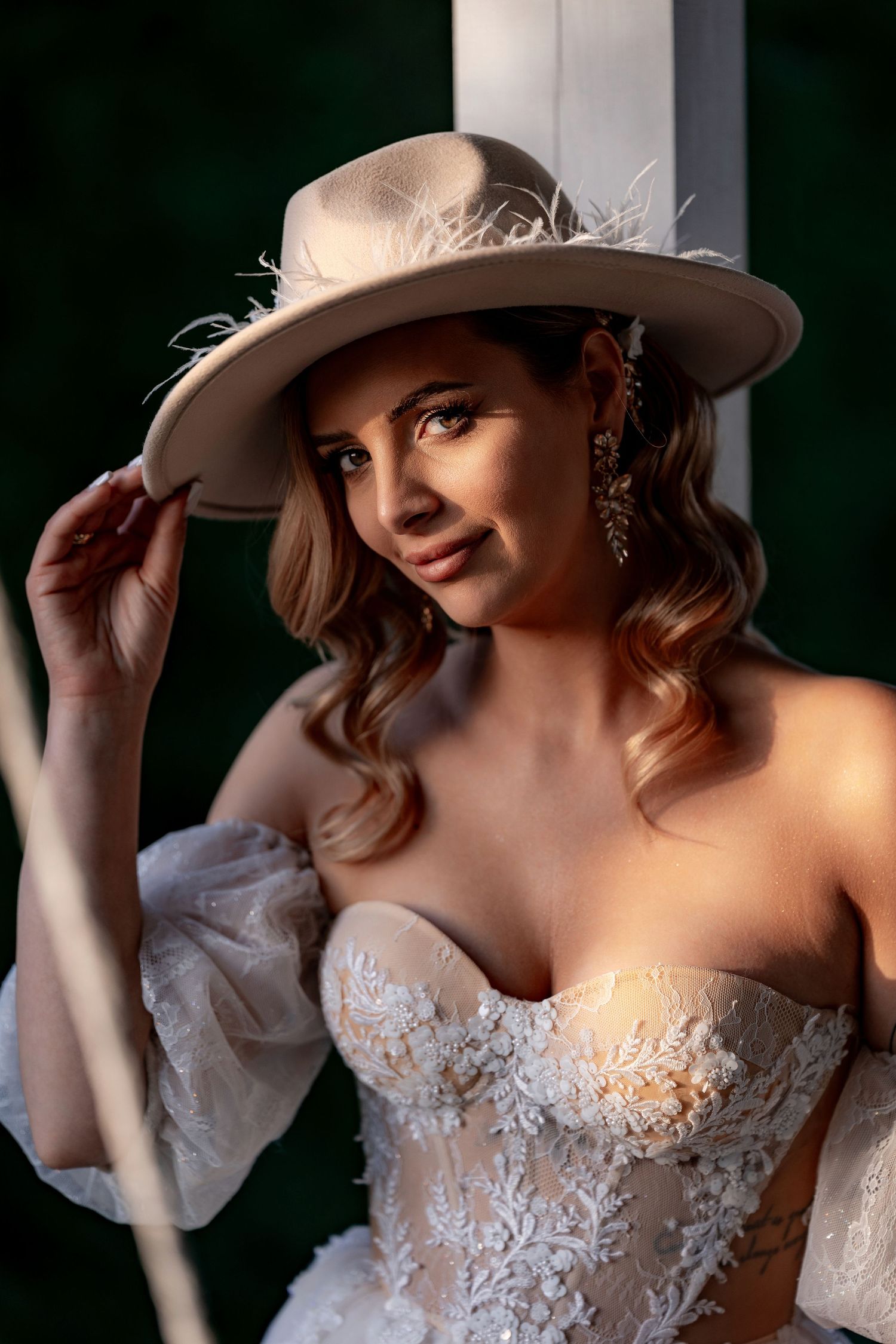 Bride in lace wedding dress poses with elegant beige hat and wavy hair.