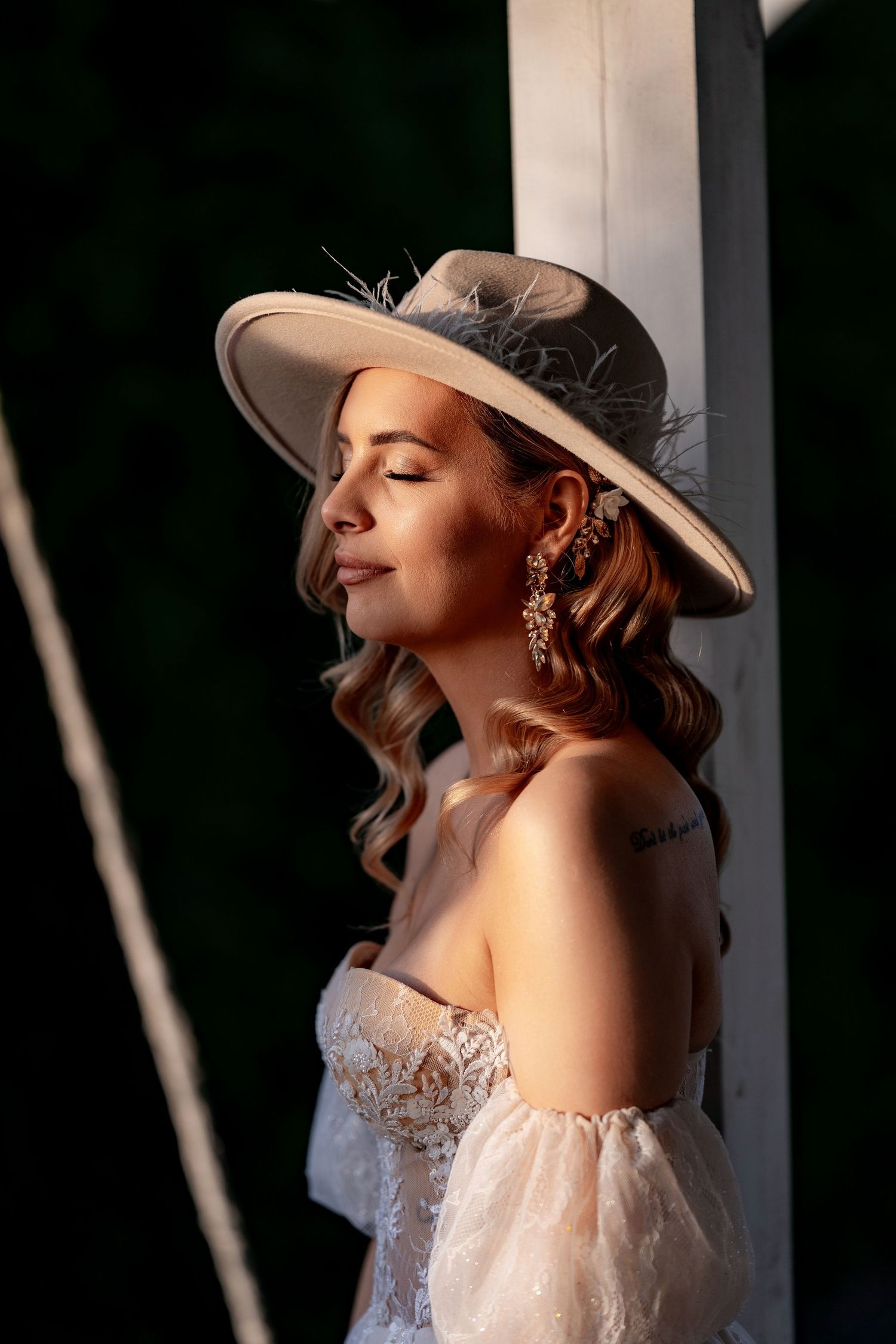 Bride in strapless gown and wide-brim hat poses in dramatic sunlight.