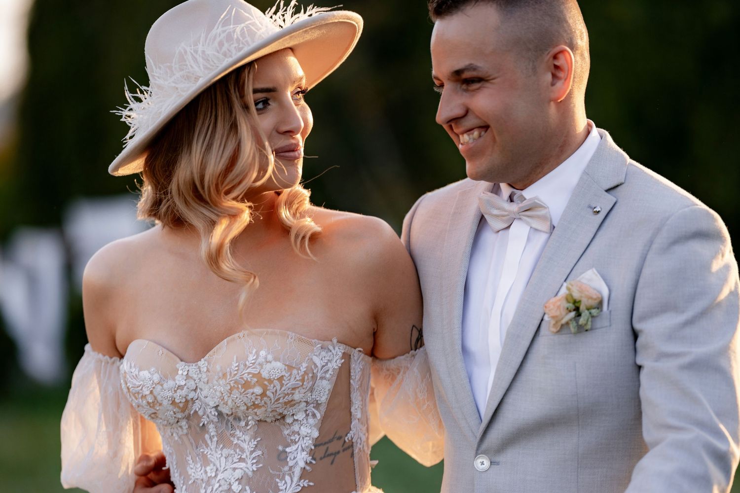 Couple shares lighthearted moment outdoors, bride in lace gown with hat and groom in light suit.