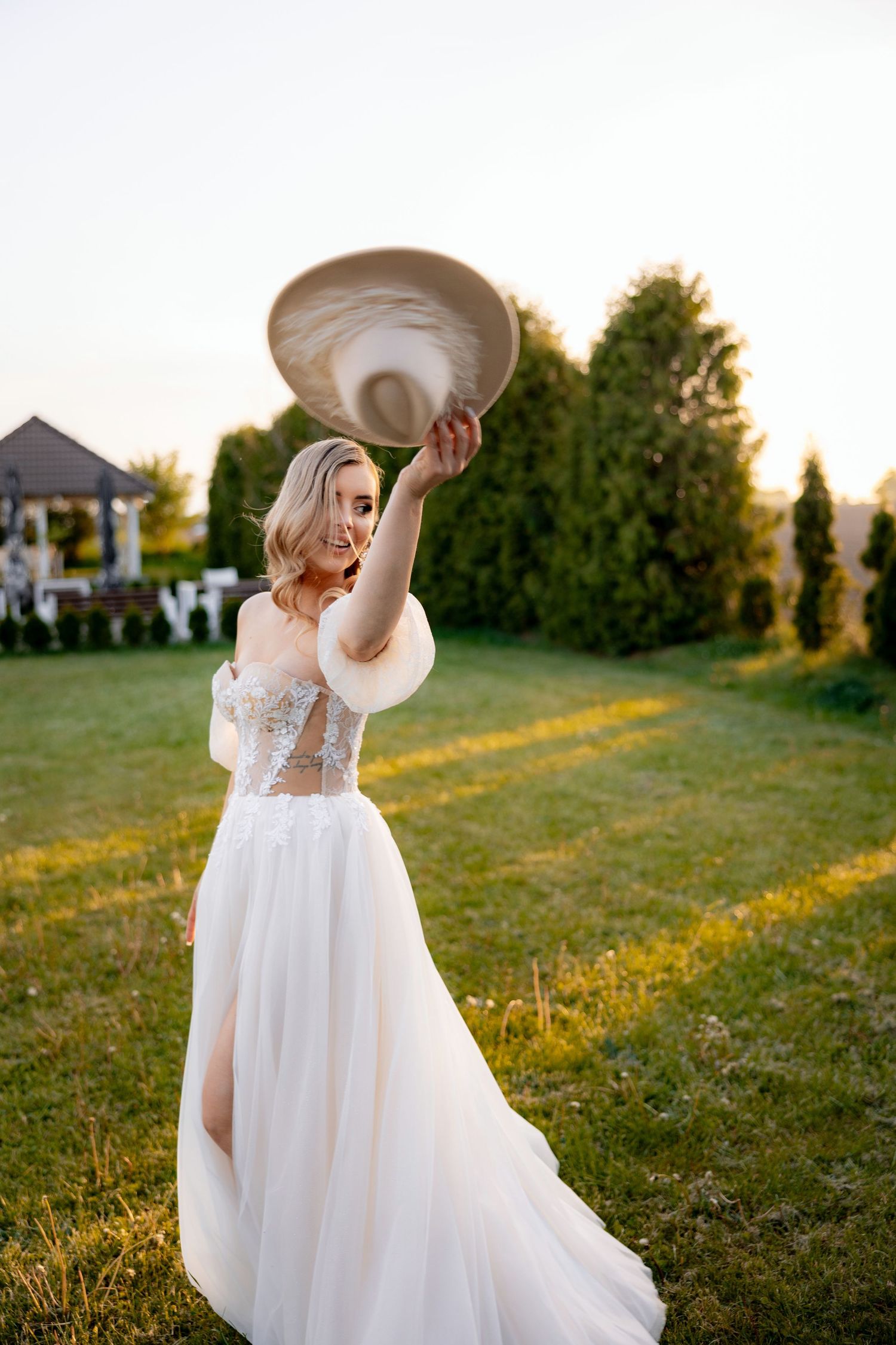 A wedding dress billows in the breeze as a light-colored hat is held aloft during sunset.