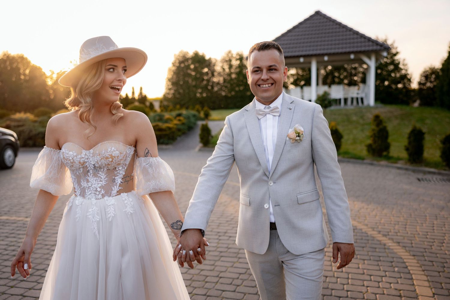 A couple in wedding attire walking hand in hand on a brick pathway at sunset.