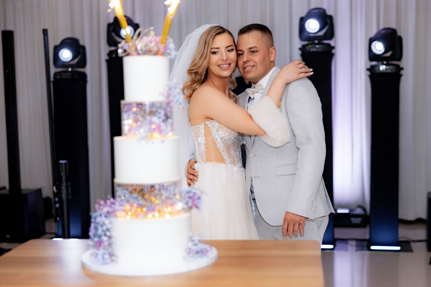 Couple embraces beside their elegant tiered wedding cake with sparkler decorations.