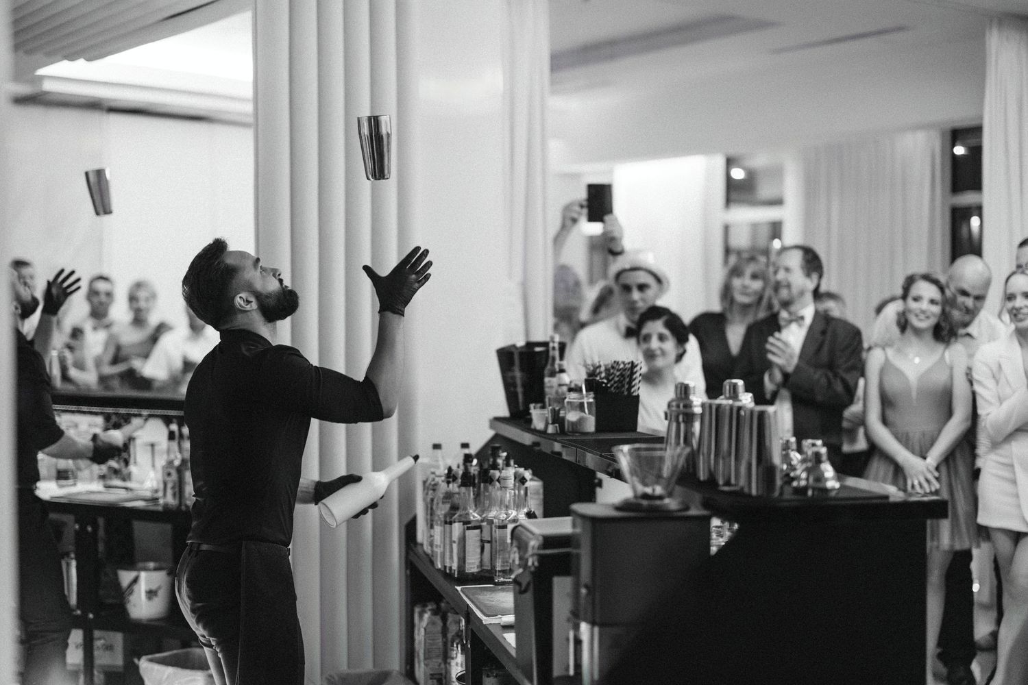 Bartender performing flair tricks behind bar while wedding guests watch and cheer.