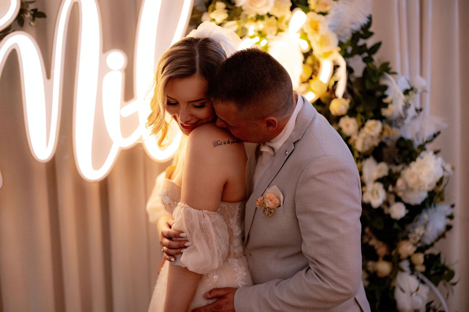 Intimate moment between newlyweds under illuminated wedding arch with white florals.