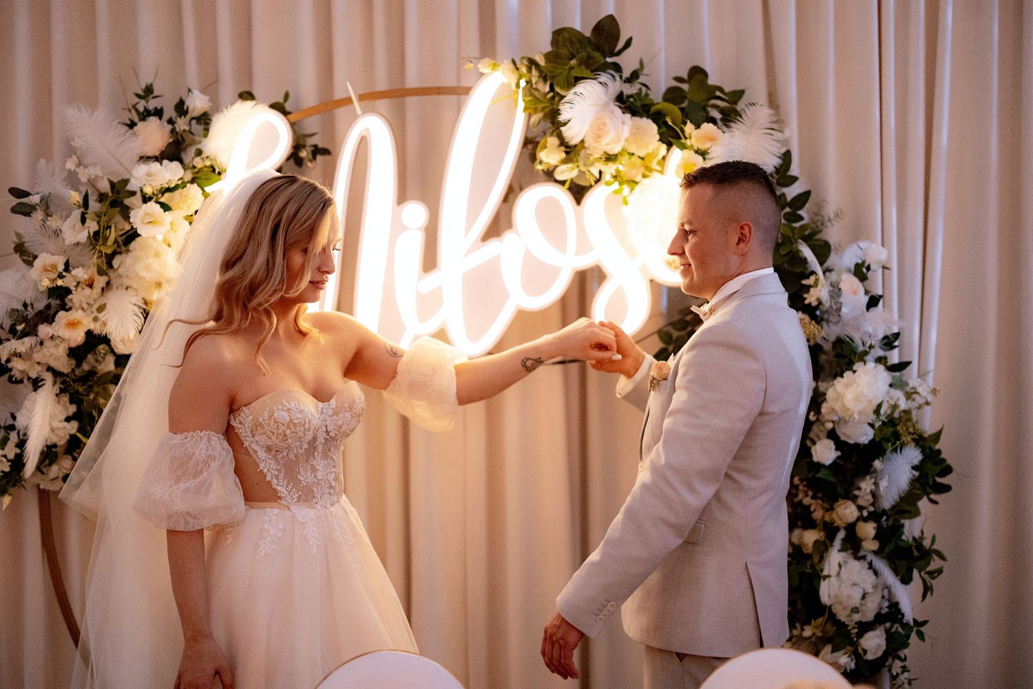 Playful wedding couple posing in front of illuminated neon sign and floral decorations.