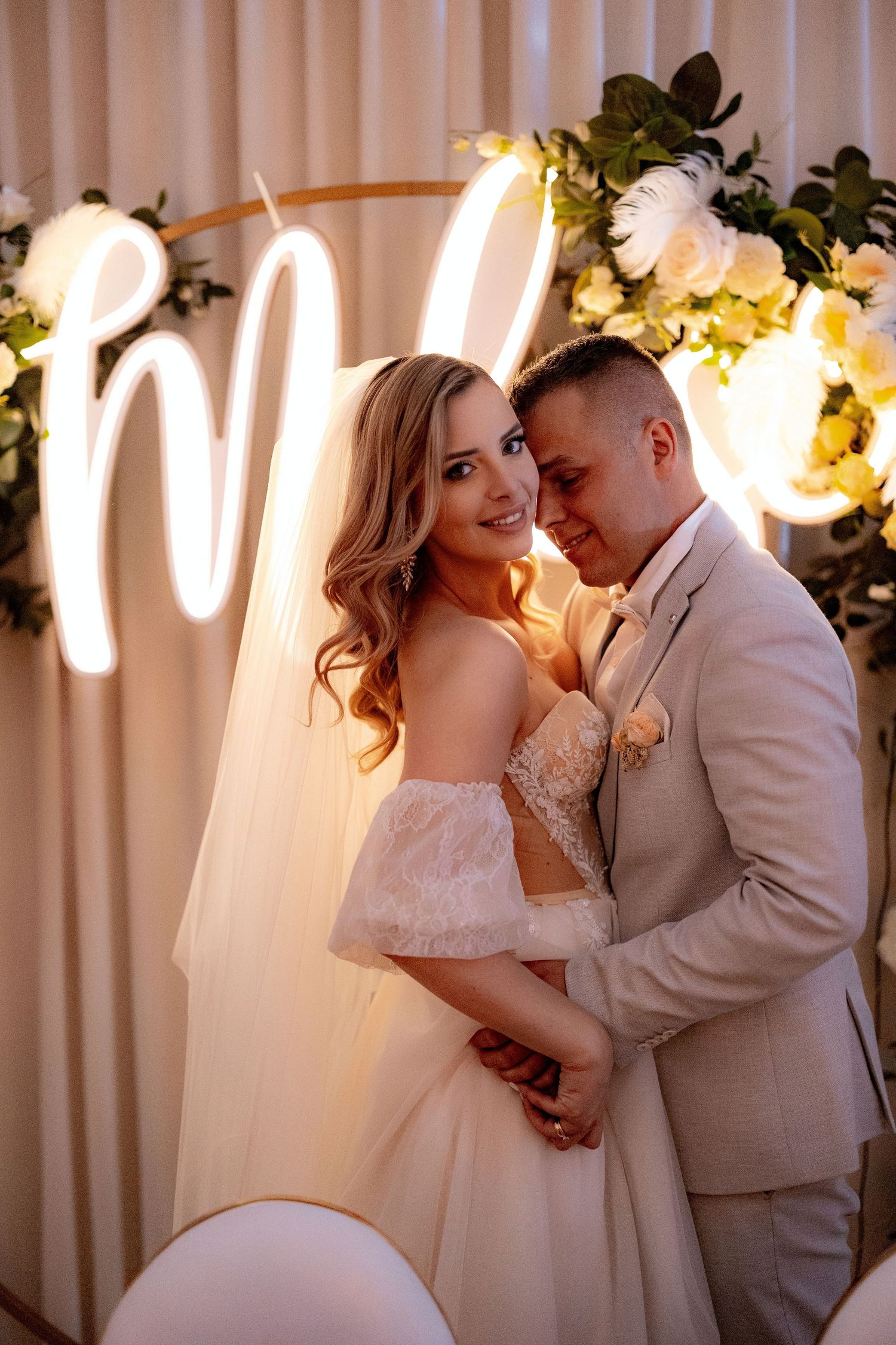 Elegant wedding portrait with couple embracing under glowing neon sign and floral arrangements.