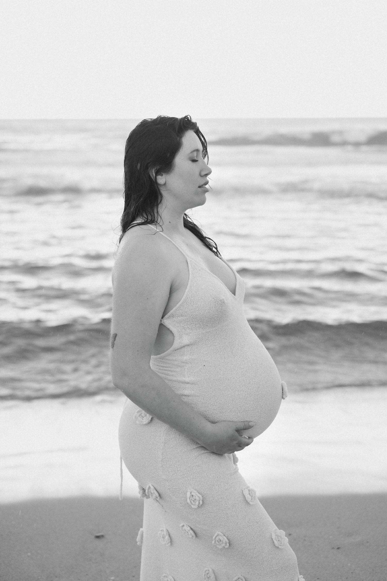 Artistic black and white maternity photoshoot series on beach with pregnant woman in long white dress.