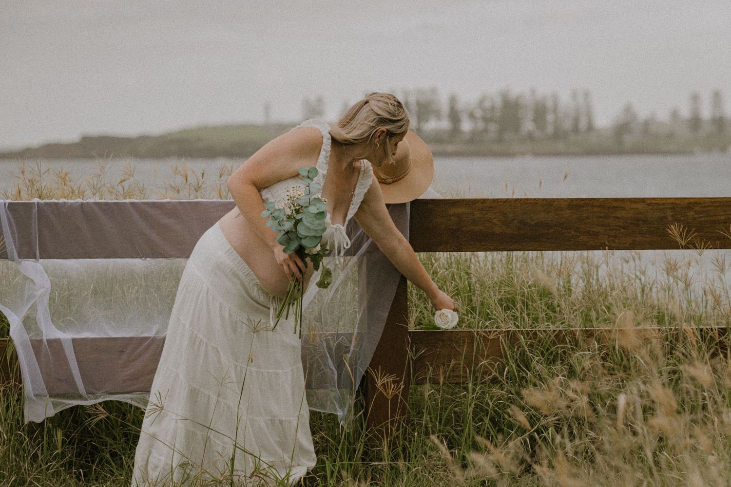 A person in a white wedding dress bends over to pick flowers in a rustic outdoor setting.