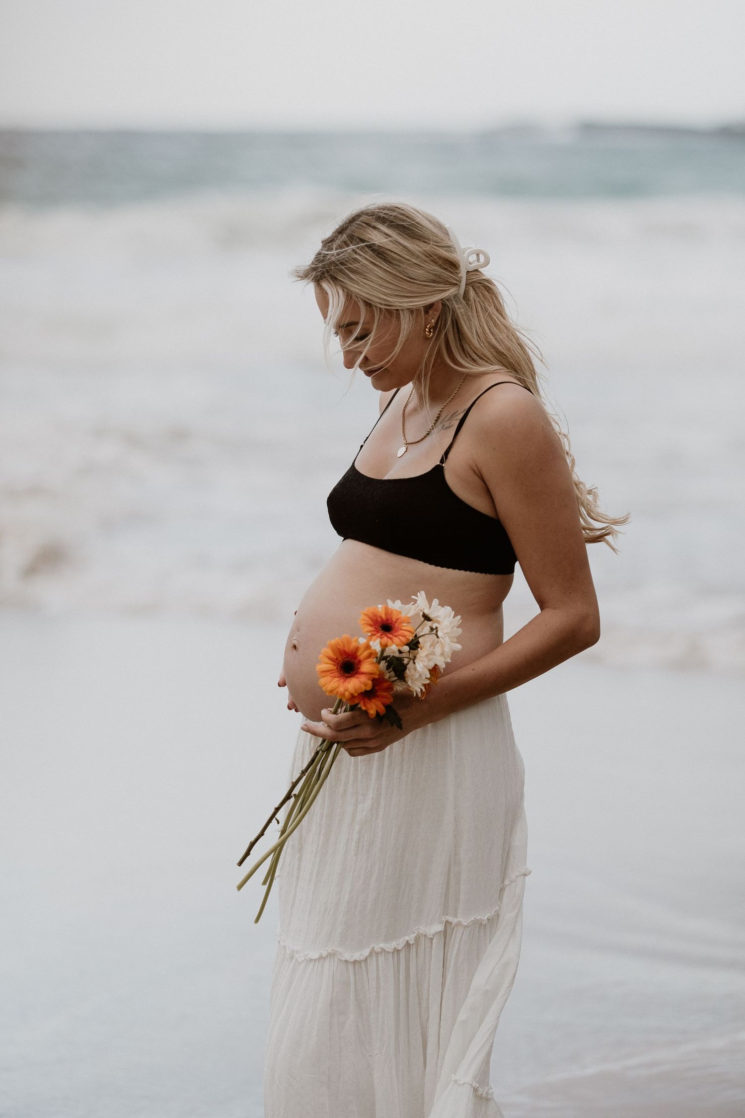 A maternity portrait on beach holding sunflowers in flowing white skirt.