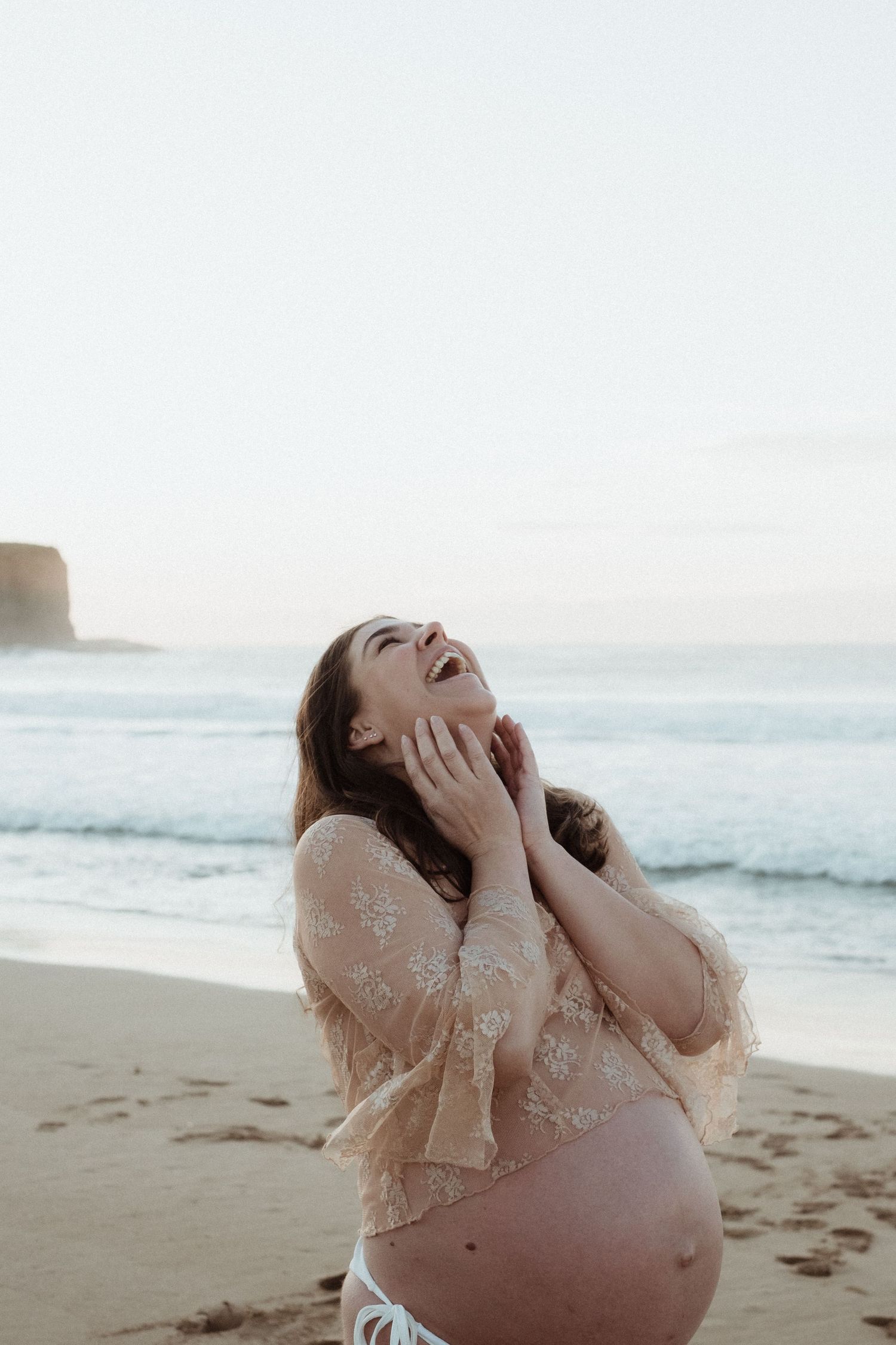 An expecting mother in a lace dress laughs joyfully on a beach during golden hour.