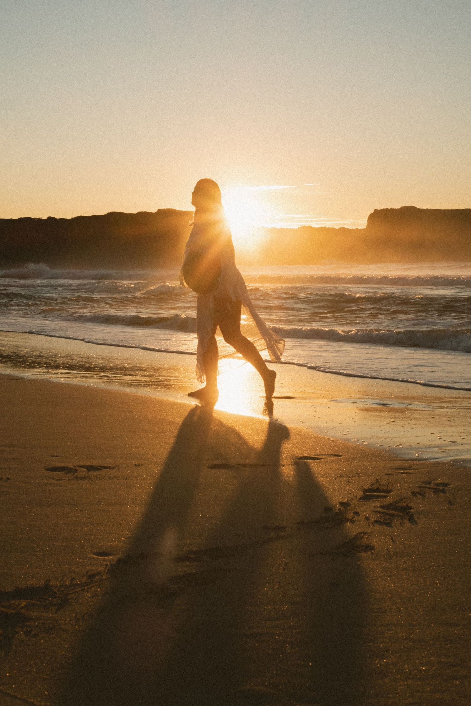 Silhouette of a person walking on a beach at sunset with waves crashing in the background.