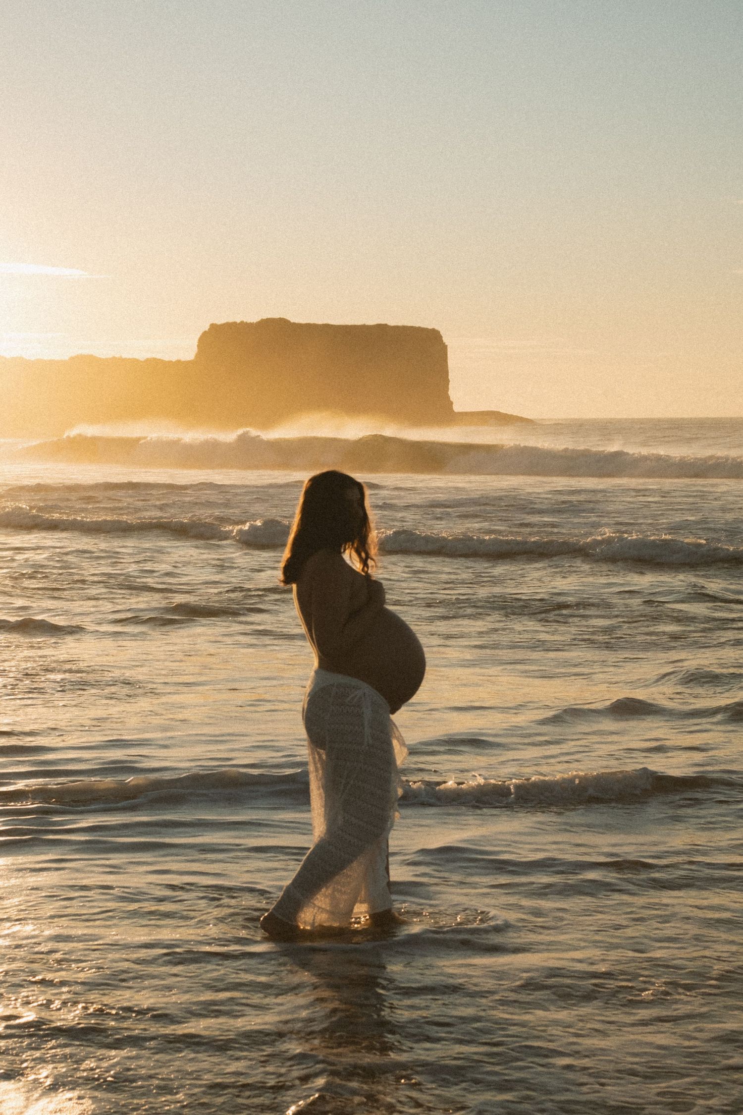 A figure in white wades into ocean waves during a dramatic coastal sunset.