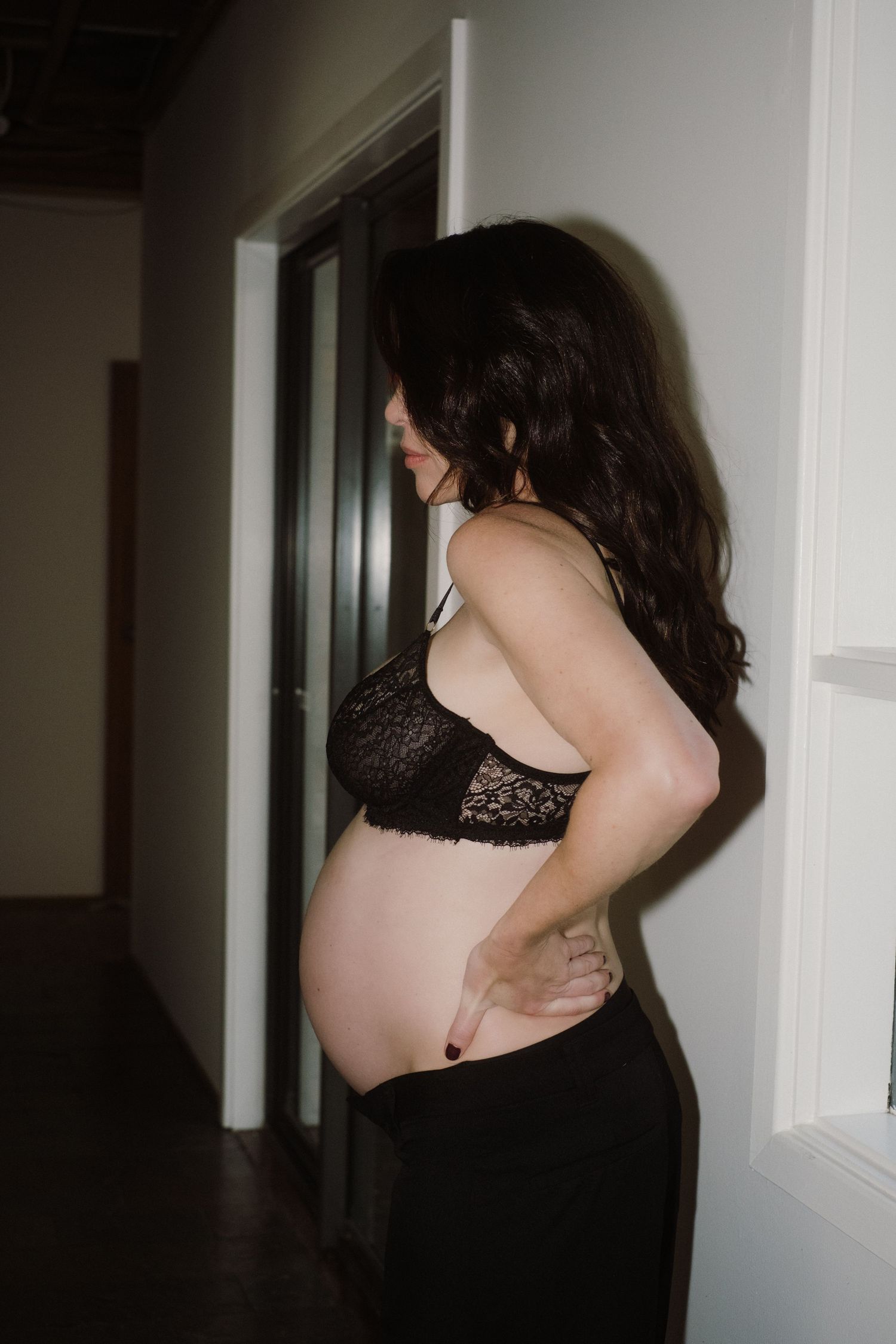 Intimate maternity photo in dimly lit hallway wearing black lace and jeans.