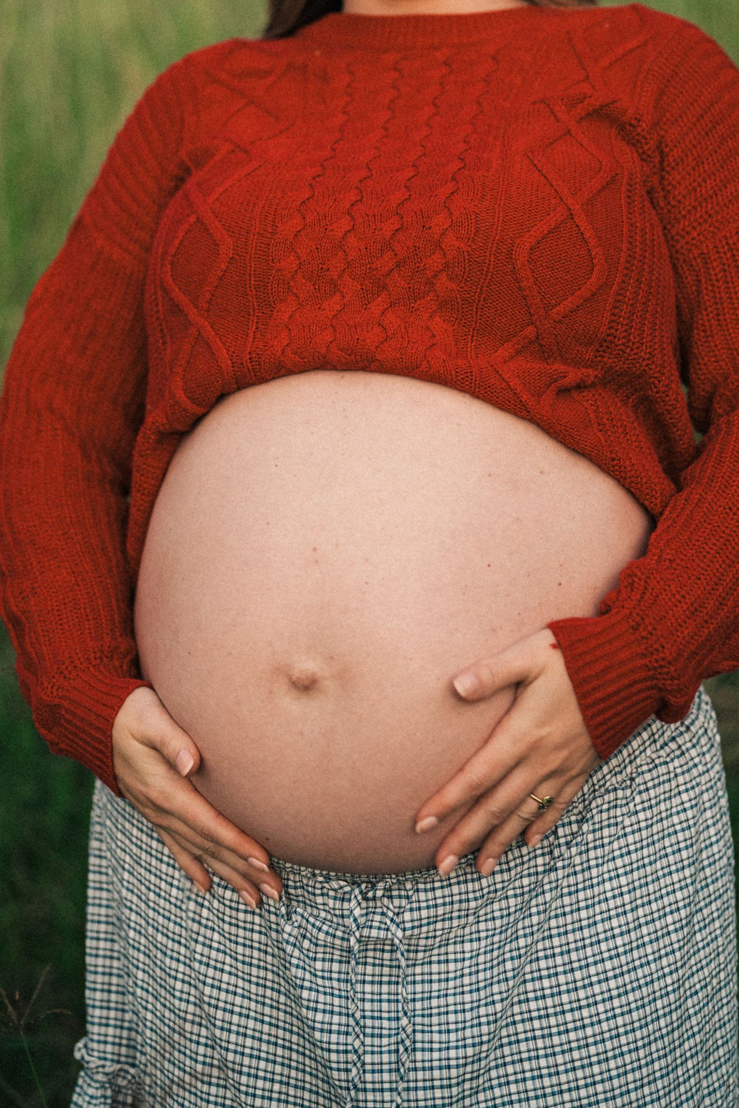 Pregnant woman in red knit sweater and plaid skirt cradling her bare baby bump outdoors.