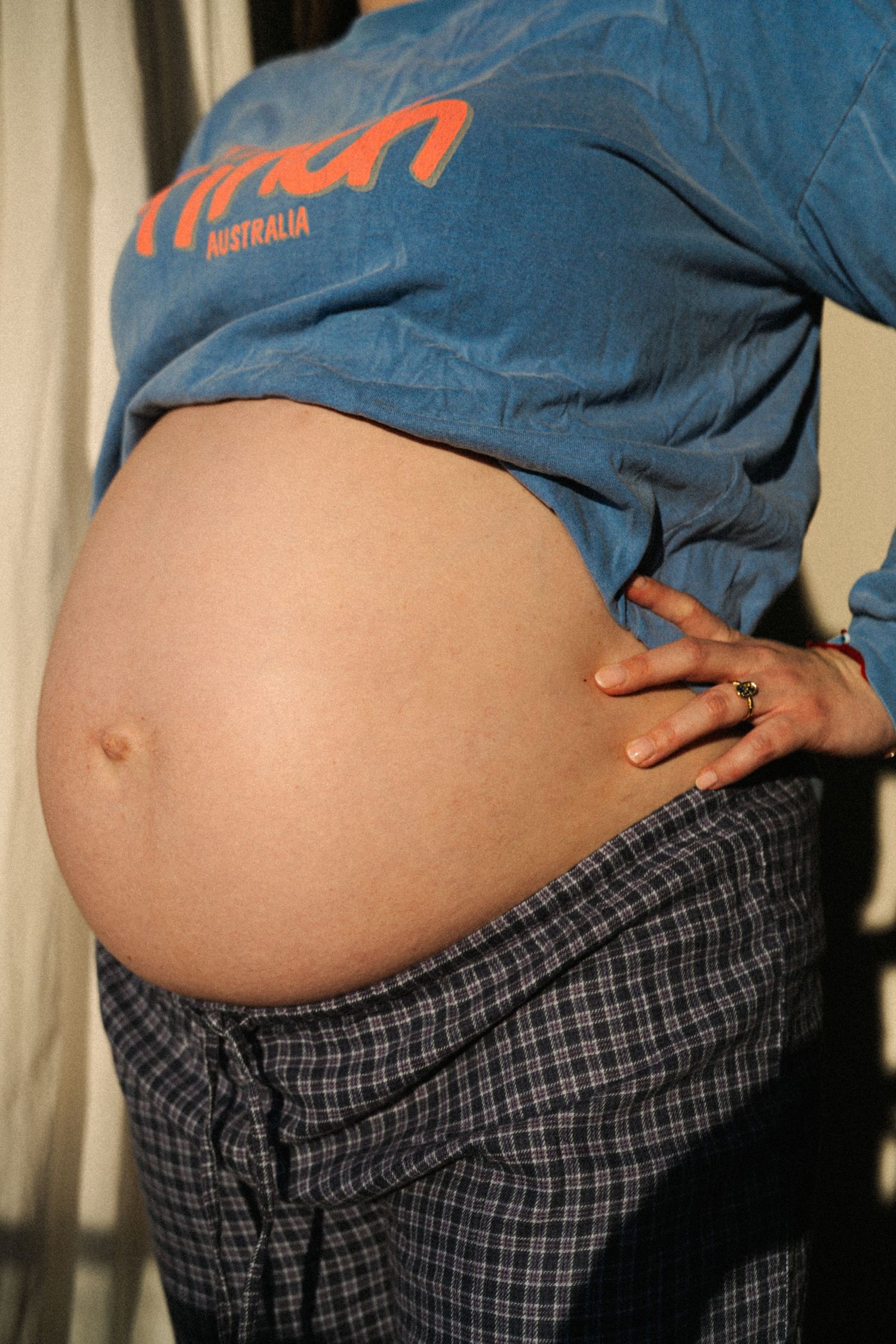 Pregnant woman in blue Australia shirt and plaid pants showing baby bump, hand on hip.