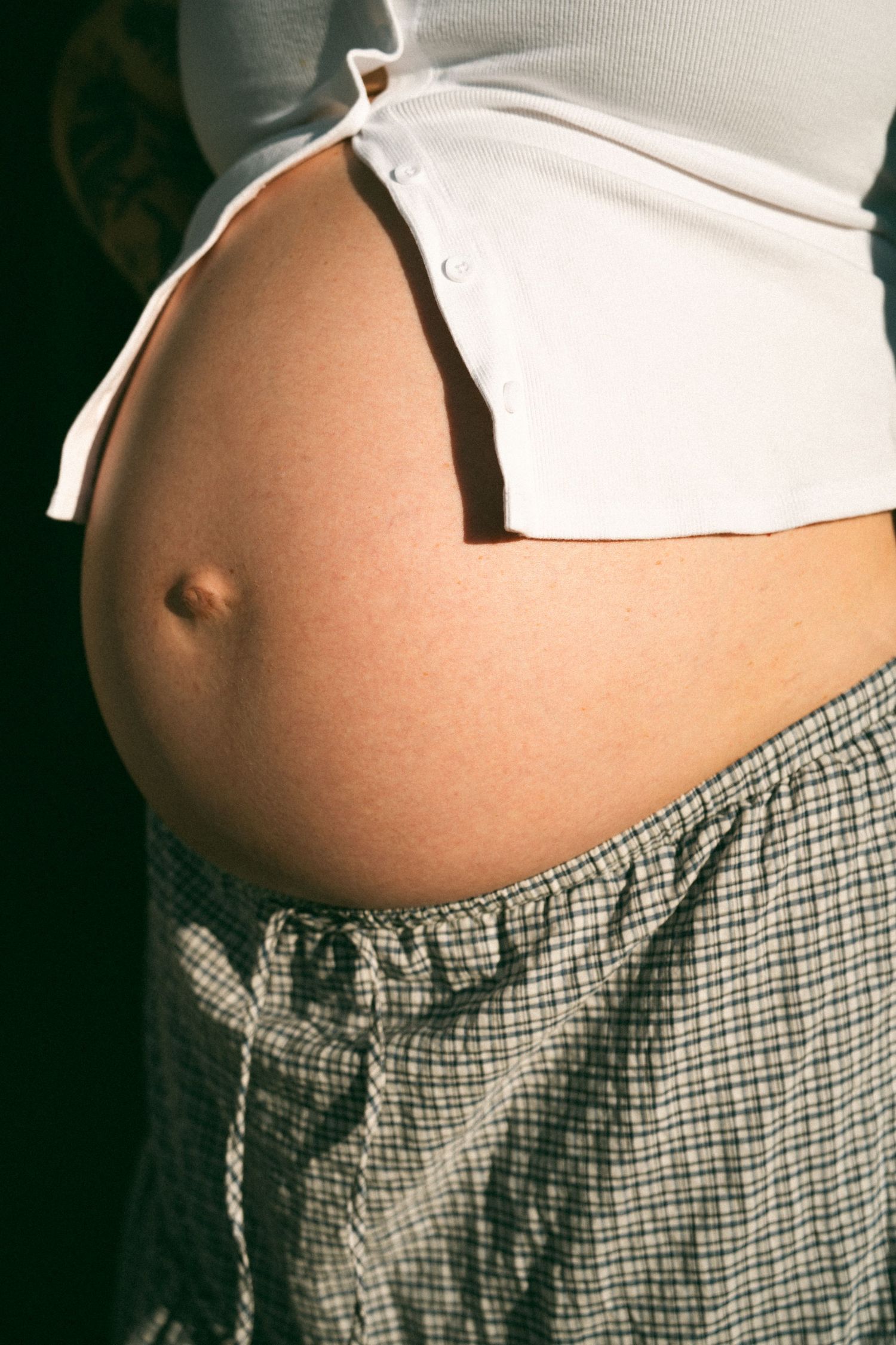 Close-up of a pregnant woman's bare baby bump in warm sunlight, wearing a white crop top and plaid skirt.