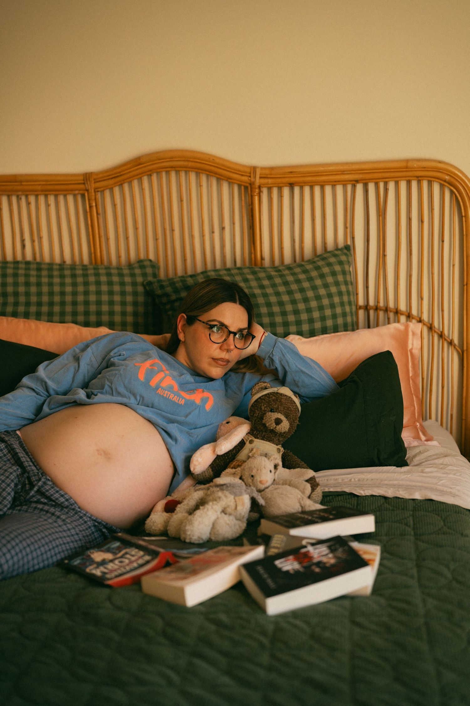 Pregnant woman relaxing on bed with teddy bears and books, wearing glasses and blue sweatshirt.