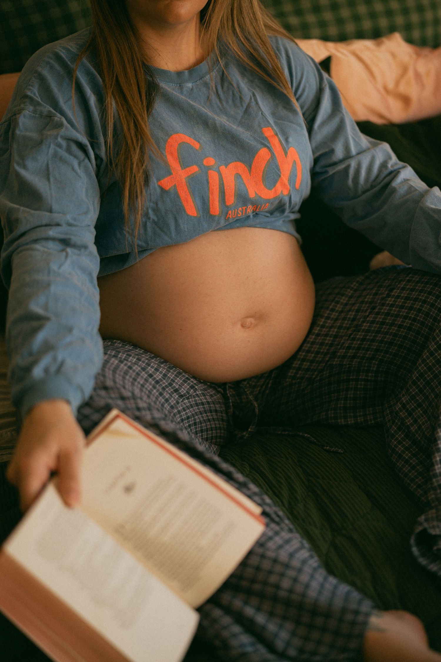 Pregnant woman in blue Finch crop top reading a book, showing baby bump while sitting comfortably.