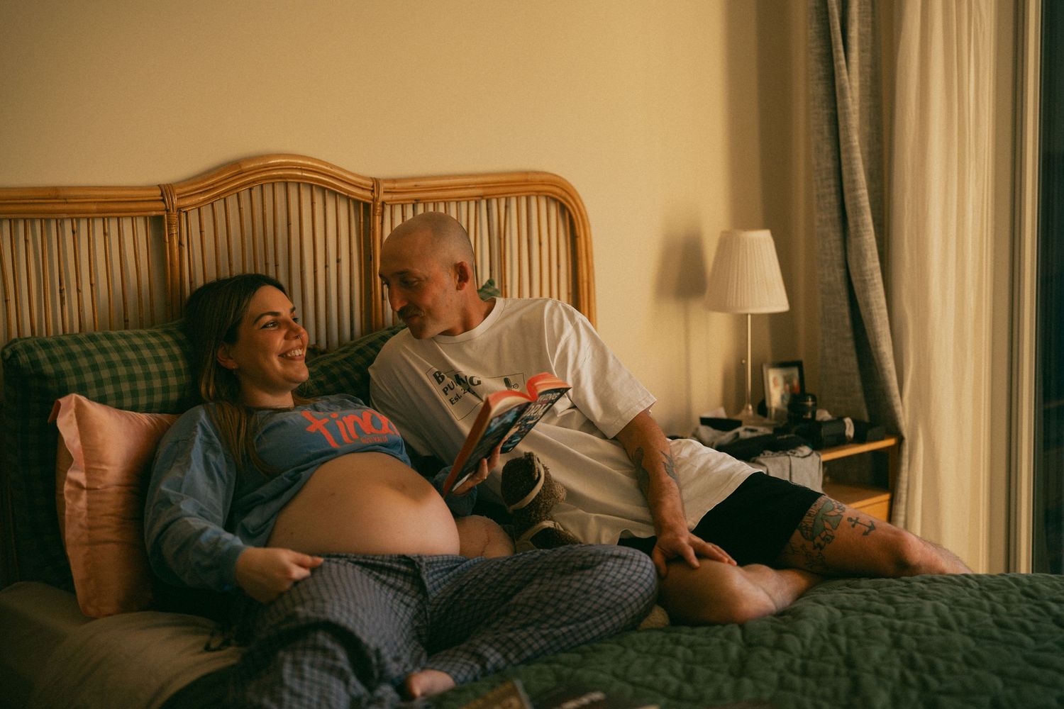 Pregnant woman and partner relaxing on bed, reading together in a cozy bedroom with warm lighting.