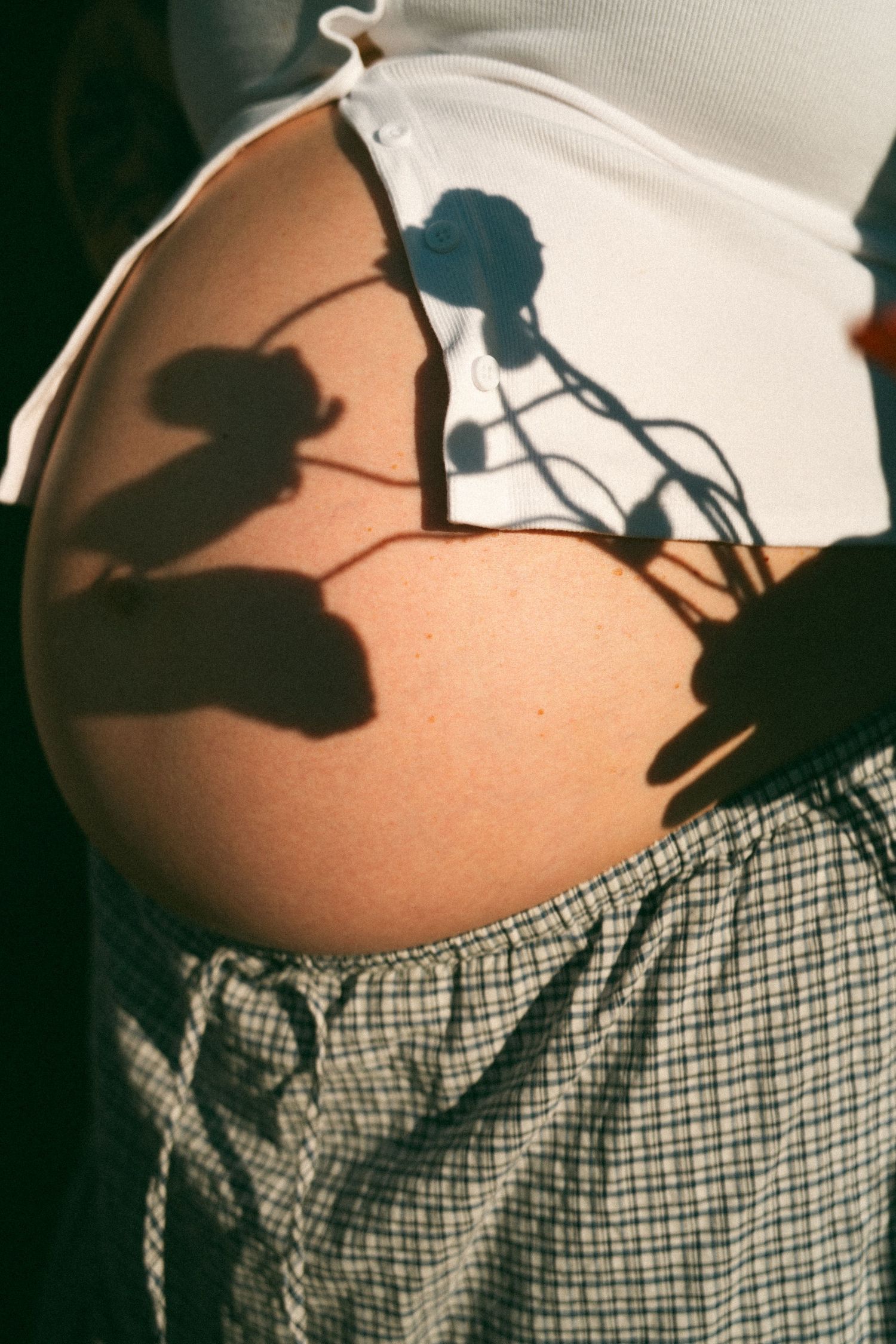 Pregnant woman's bare baby bump with floral shadows cast by sunlight, wearing a white crop top and plaid skirt.