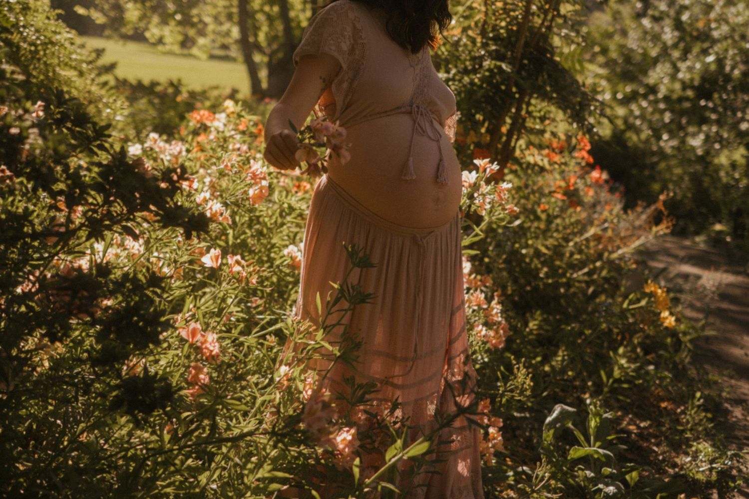 Person in a flowy pink dress stands in a sunlit garden surrounded by blooming flowers and greenery.