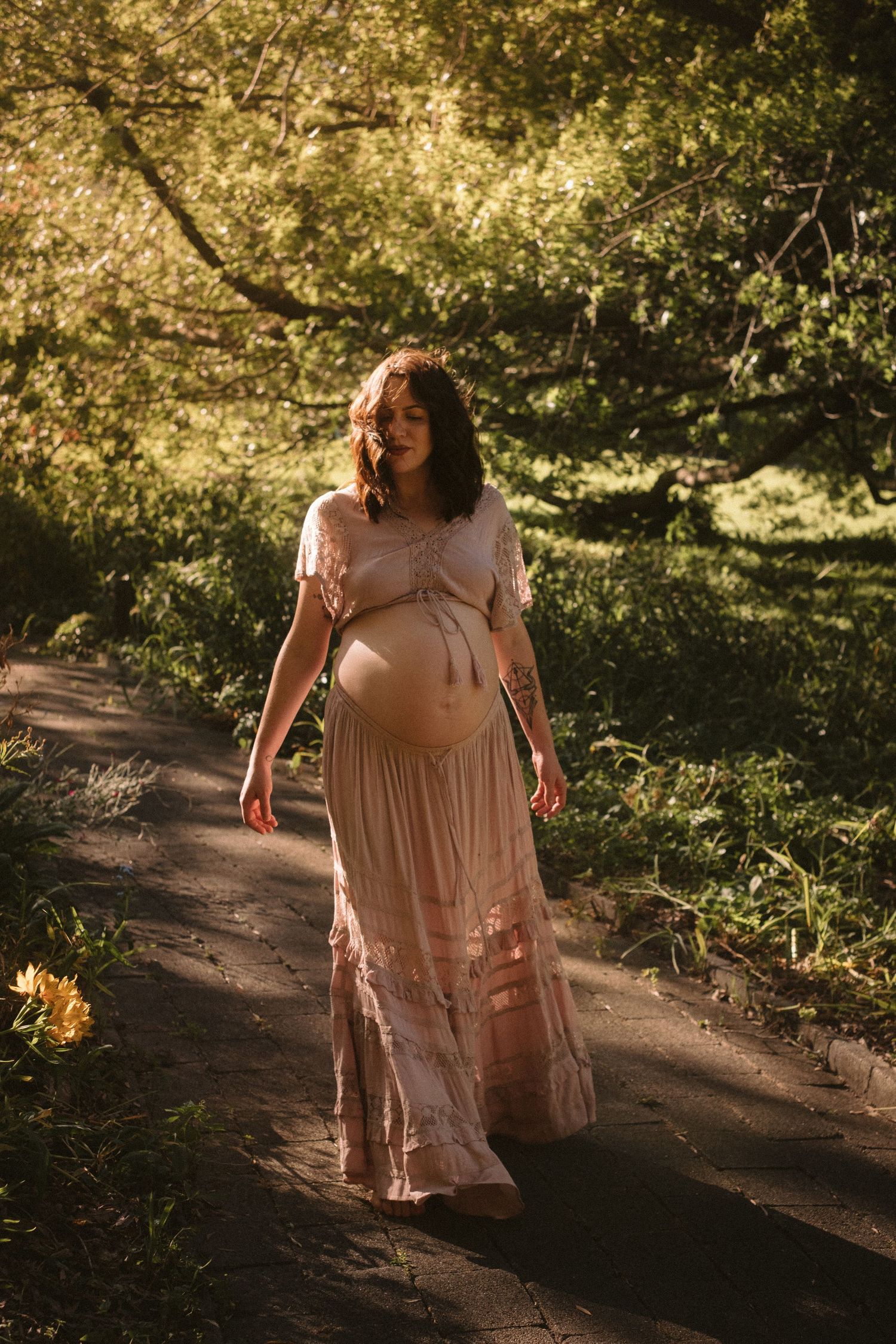 A person in a flowing beige dress walks along a sunlit dirt path through a wooded area in a dreamy outdoor setting.