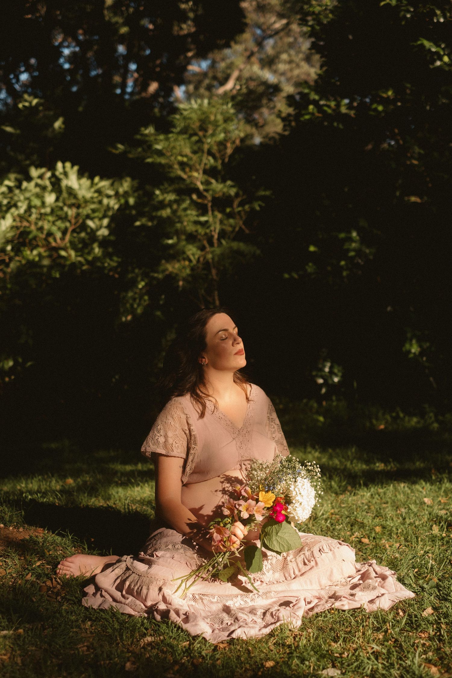 A person in a pink dress reclines on grass in dappled sunlight while holding a bouquet of flowers.