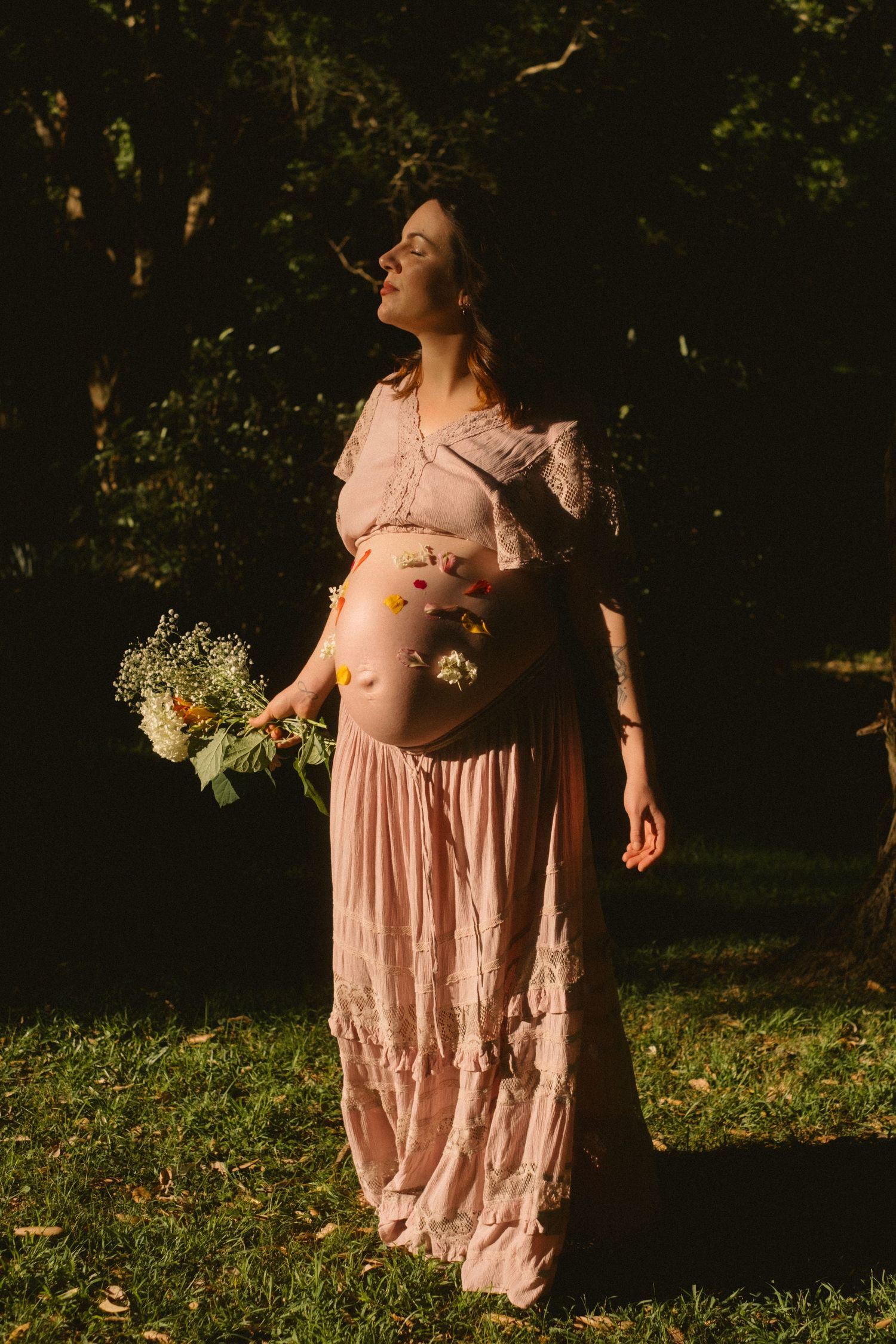 A figure in a flowing pink dress stands in dappled sunlight among trees in a dreamy outdoor setting.