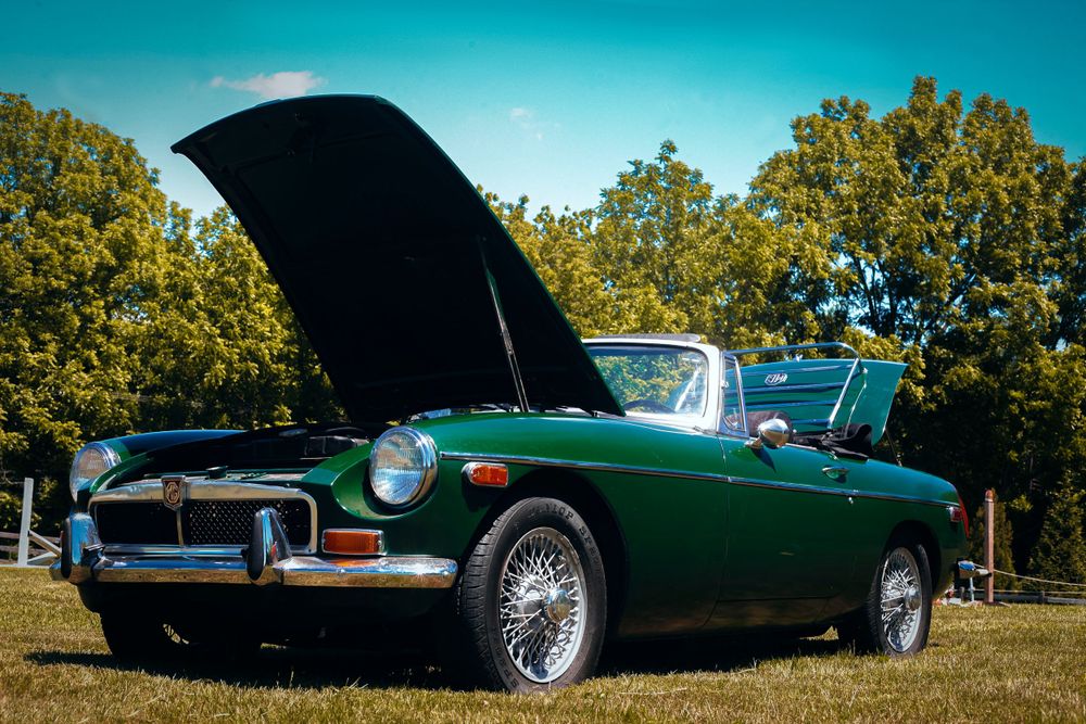 Green MG convertible sports car parked on grass against blue sky with trees in background.