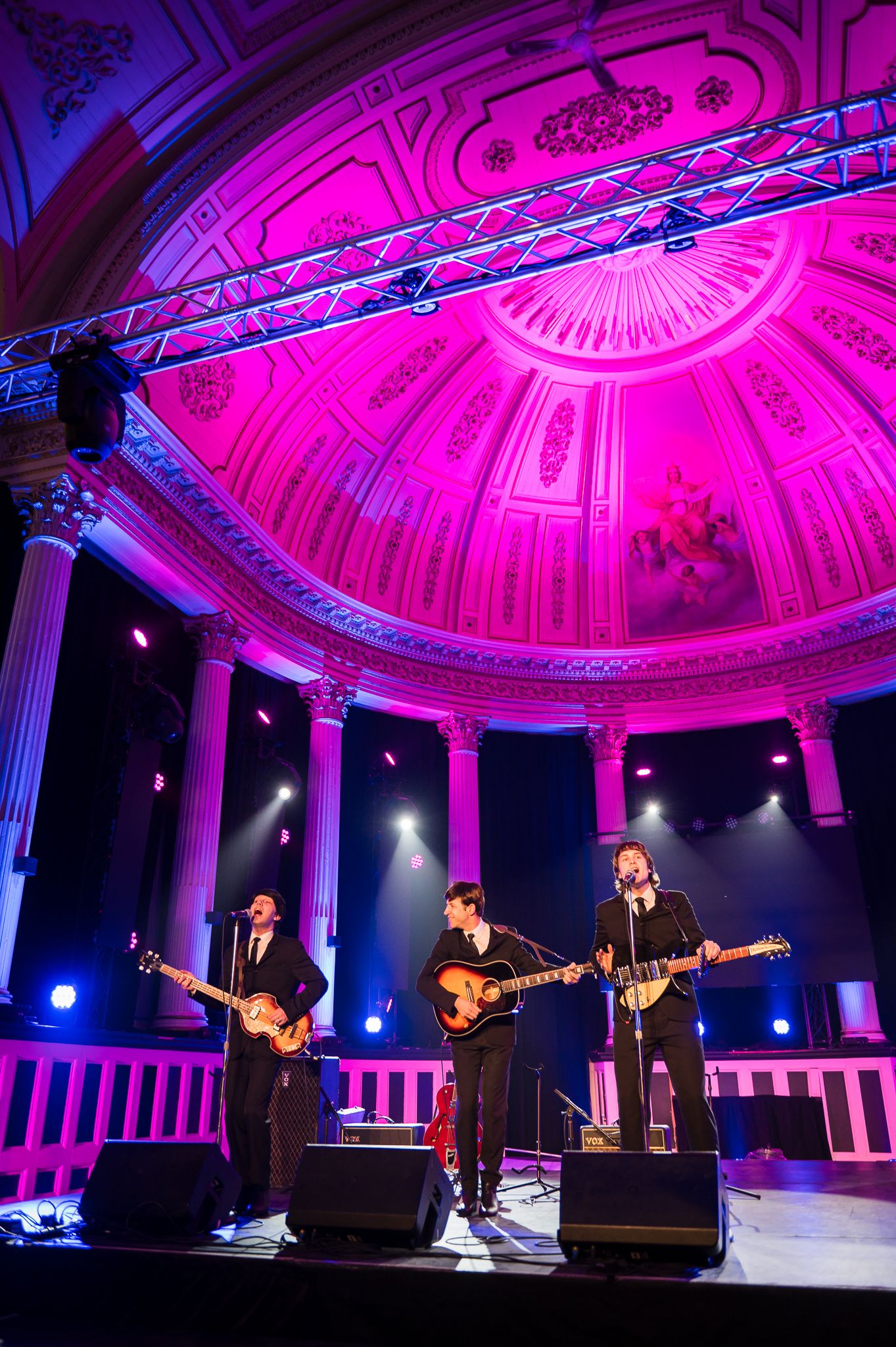 Band performing on stage under dramatic pink-lit dome with ornate architectural details.