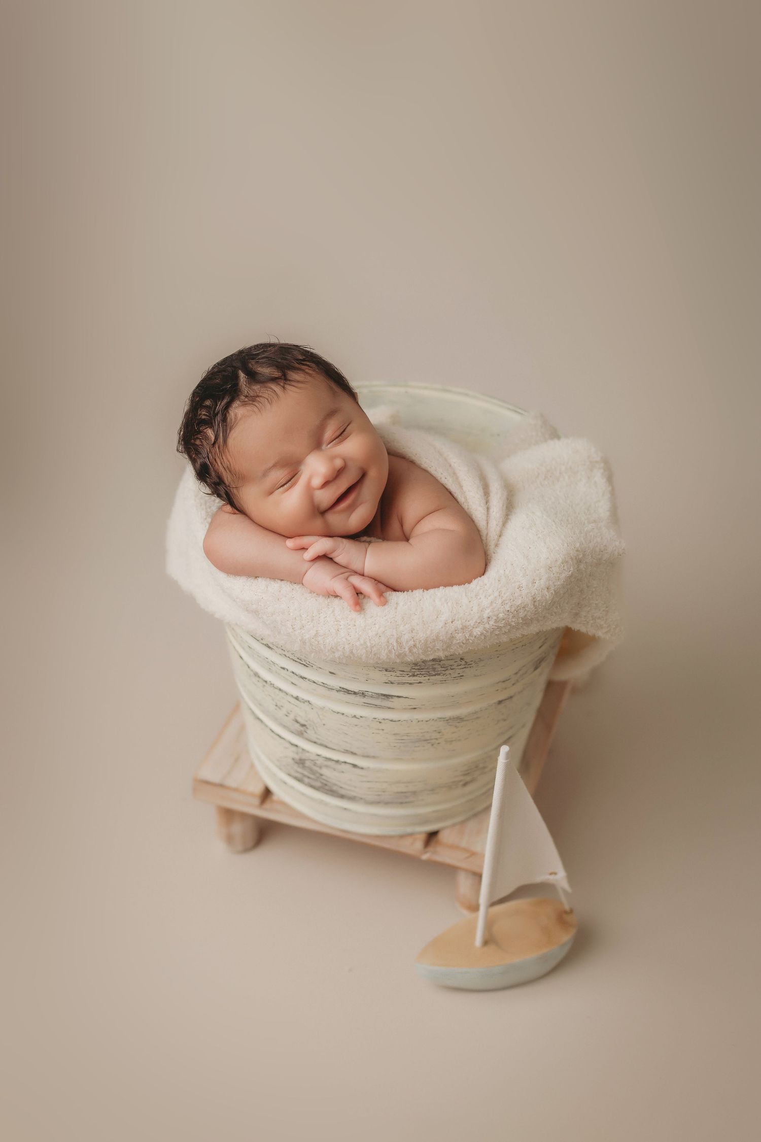 Sleeping newborn wrapped in white fabric in small wooden prop with tiny sailboat.