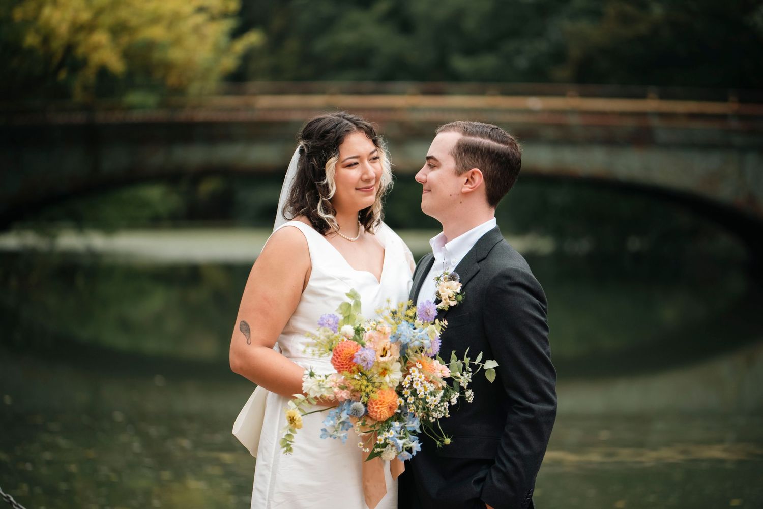 Newlywed couple sharing intimate moment by bridge with autumn bouquet.