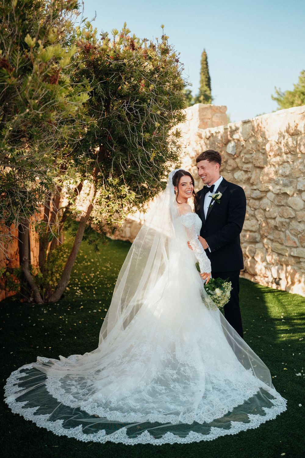 Bride in lace gown with long veil and groom in black tuxedo posing together in a garden with stone wall.