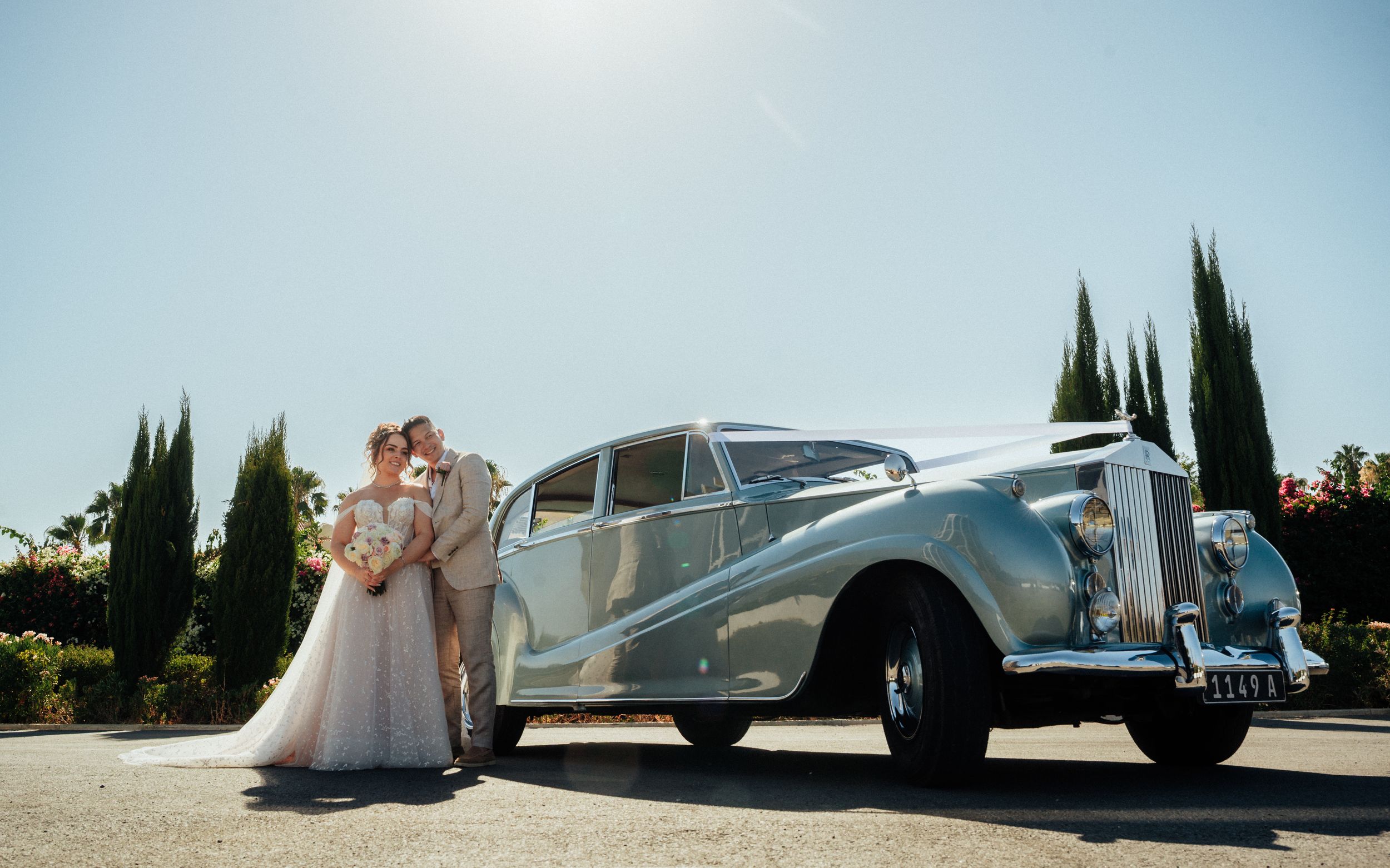 Bride and groom posing beside a classic vintage white Rolls-Royce wedding car on a sunny day.