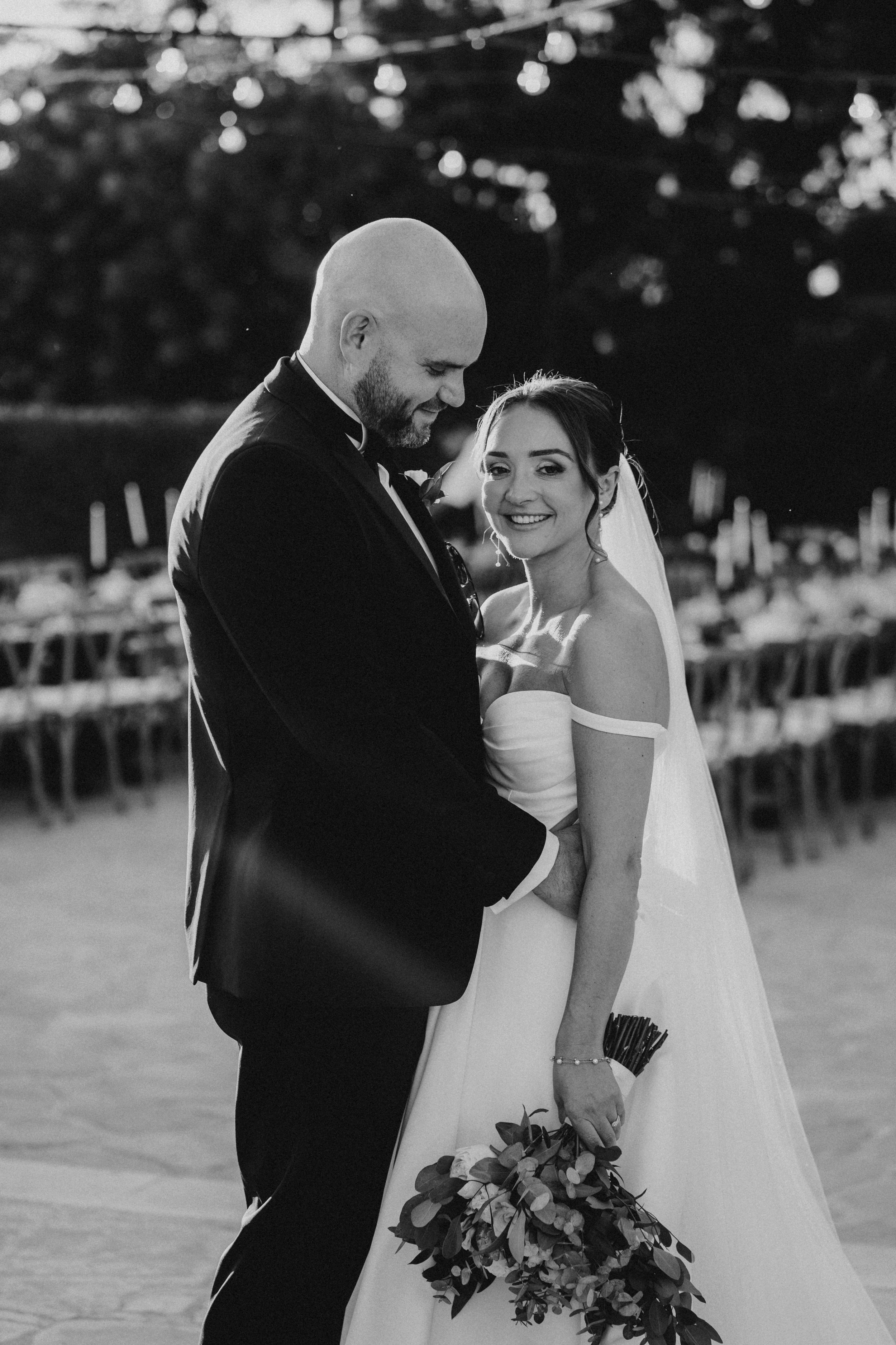 Black and white wedding photo of a couple sharing a joyful moment outdoors with twinkling lights and reception seating visible.