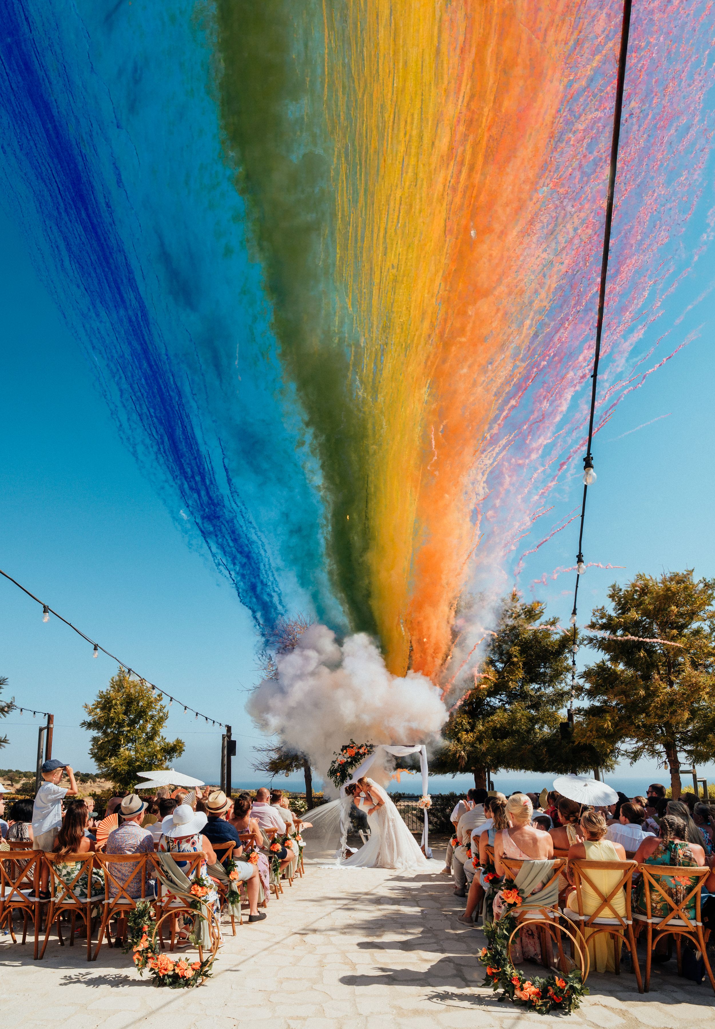 Outdoor wedding ceremony with colorful rainbow smoke cannons creating a vibrant arc over the couple kissing at the altar.