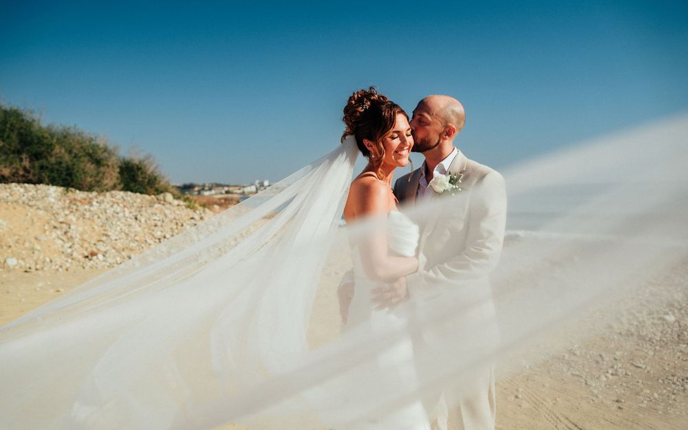 Bride and groom share a romantic moment on a sandy beach, her flowing veil billowing in the breeze under a clear blue sky.