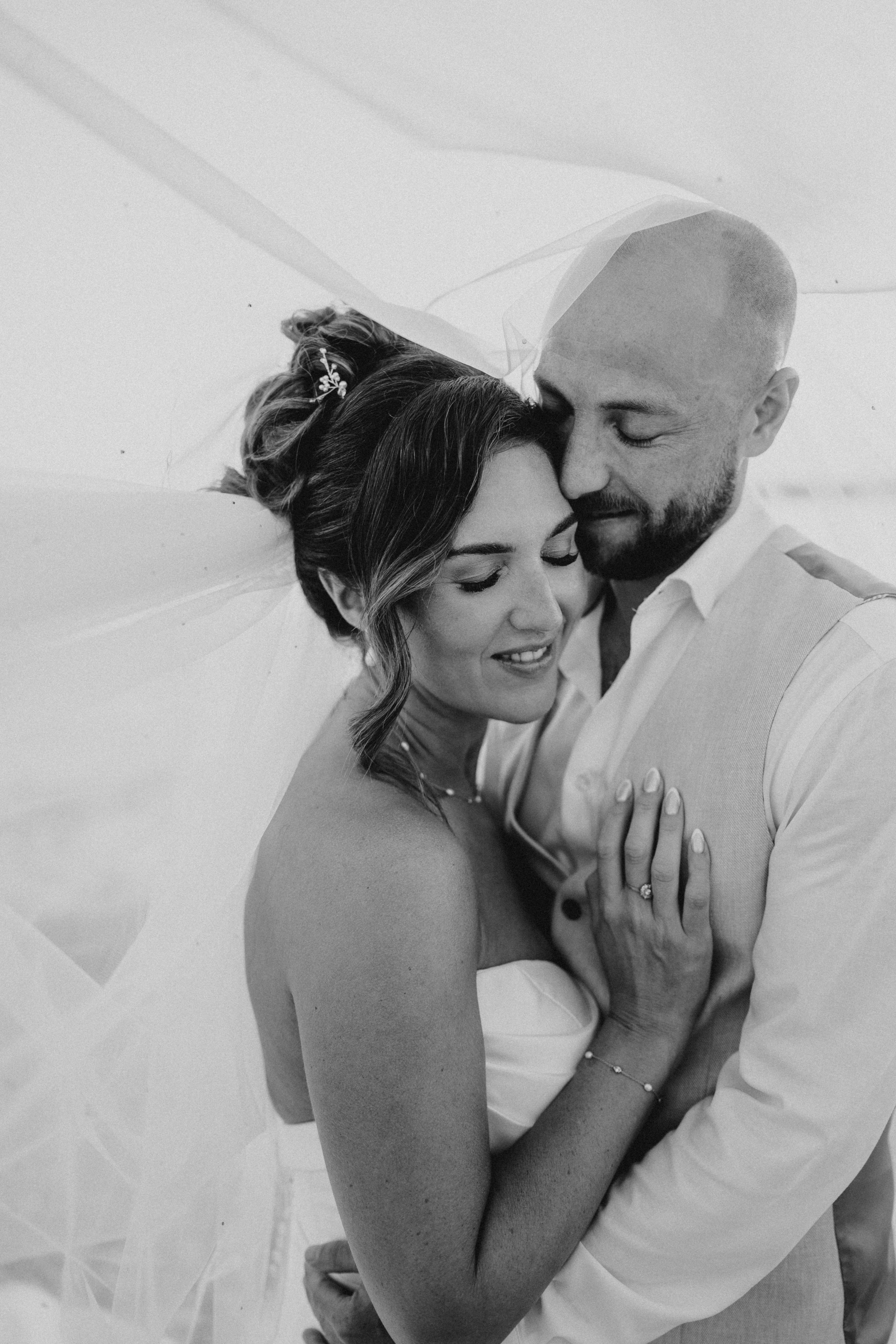 Black and white wedding photo of bride and groom embracing tenderly under a flowing veil.