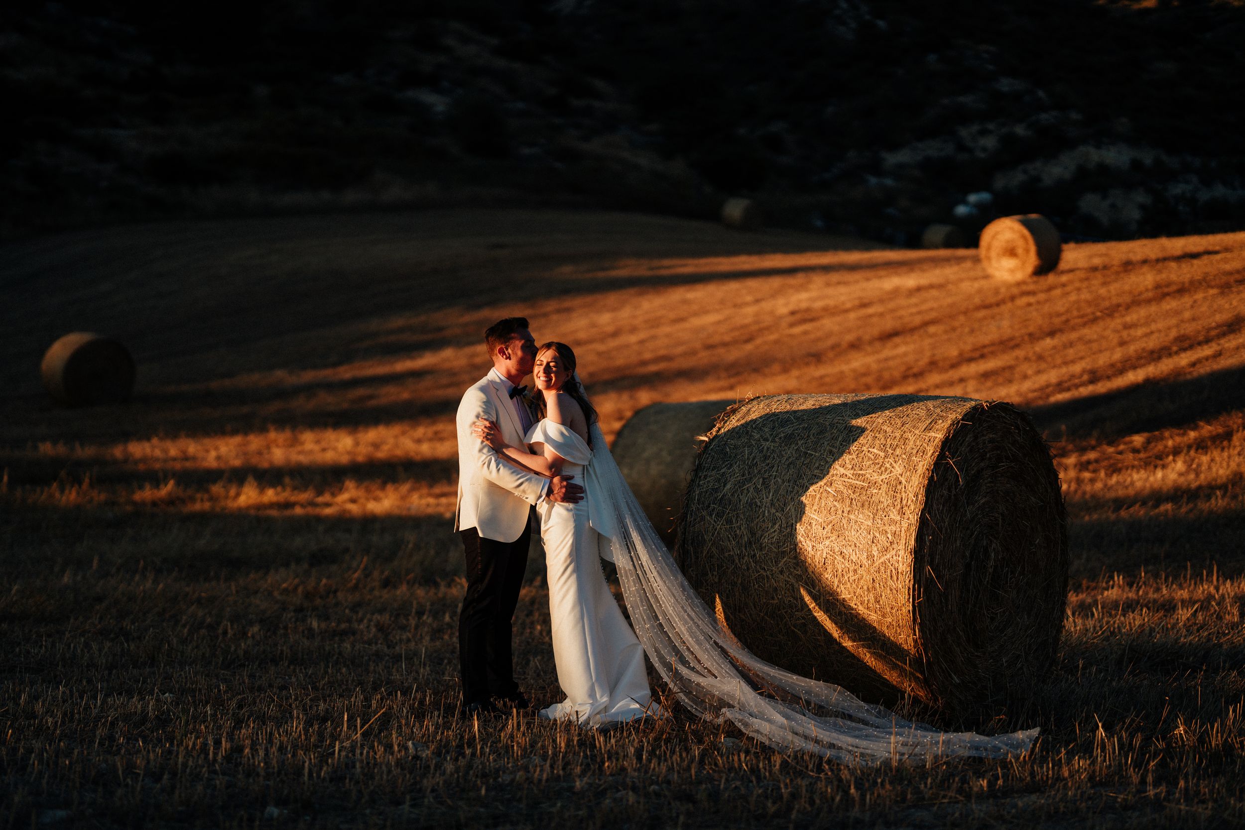 Bride and groom embracing at golden hour beside hay bales in a sunlit field, bride wearing a flowing white gown with long veil.